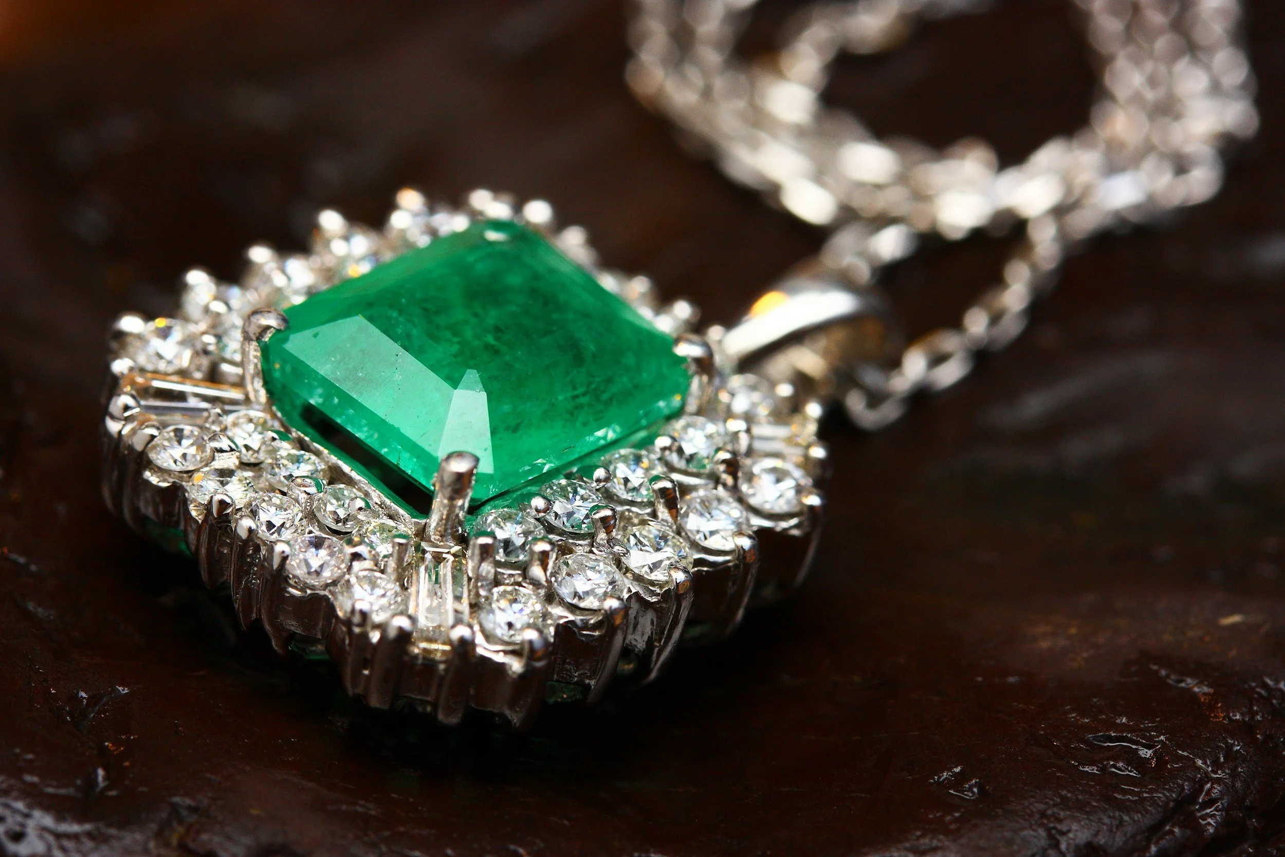 A silver necklace with a large green gemstone surrounded by small clear diamonds on a dark surface.