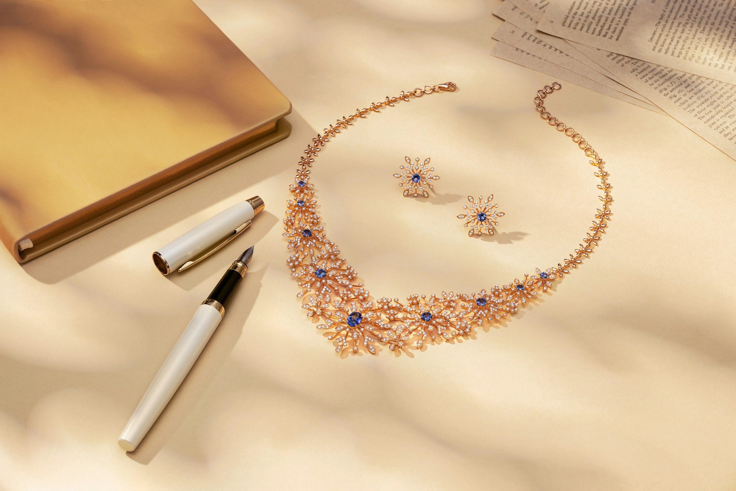 Gold jewelry set including a necklace and earrings with blue gemstones and diamonds, placed on a white surface with a notebook, papers, and pens nearby.