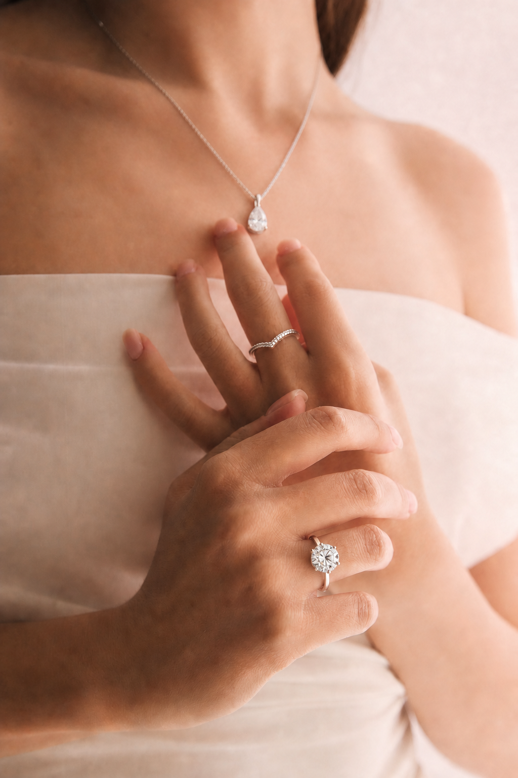 Close-up of a woman’s hands and upper chest, showing a diamond engagement ring on her finger and two rings, as well as a necklace with a teardrop-shaped diamond pendant.