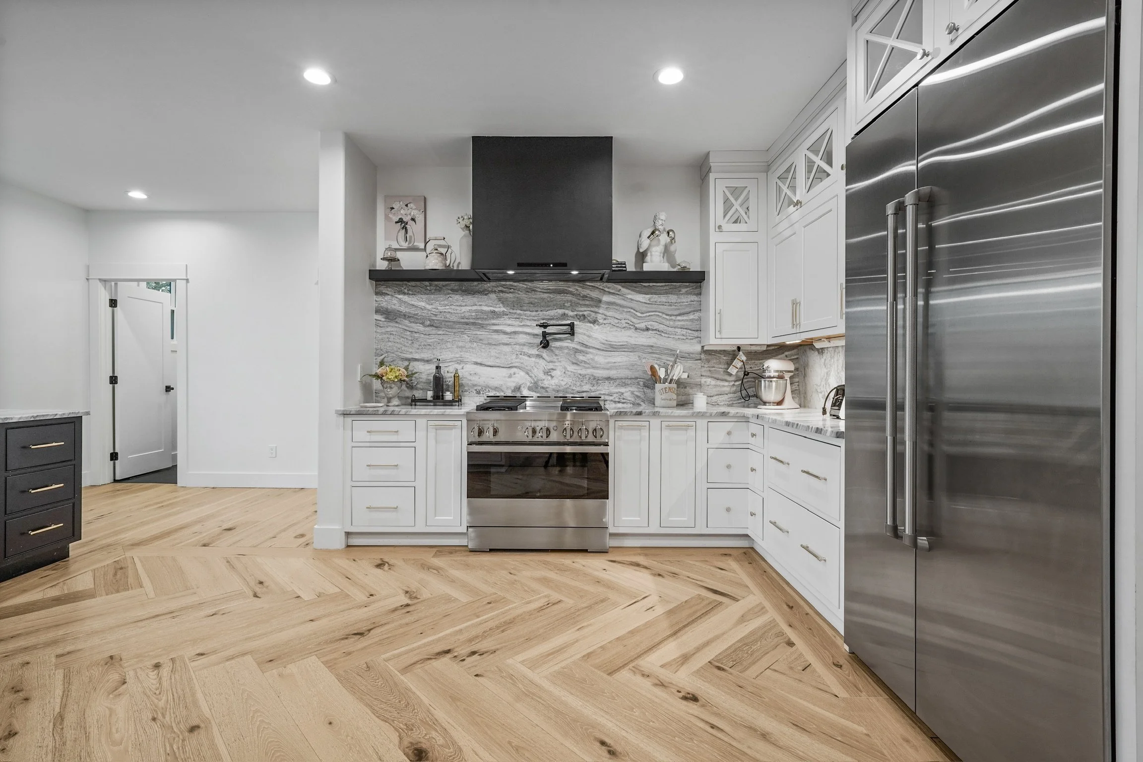 Modern kitchen with white cabinets, marble backsplash, stainless steel appliances, light wood flooring, and decorative ornaments on a black shelf.