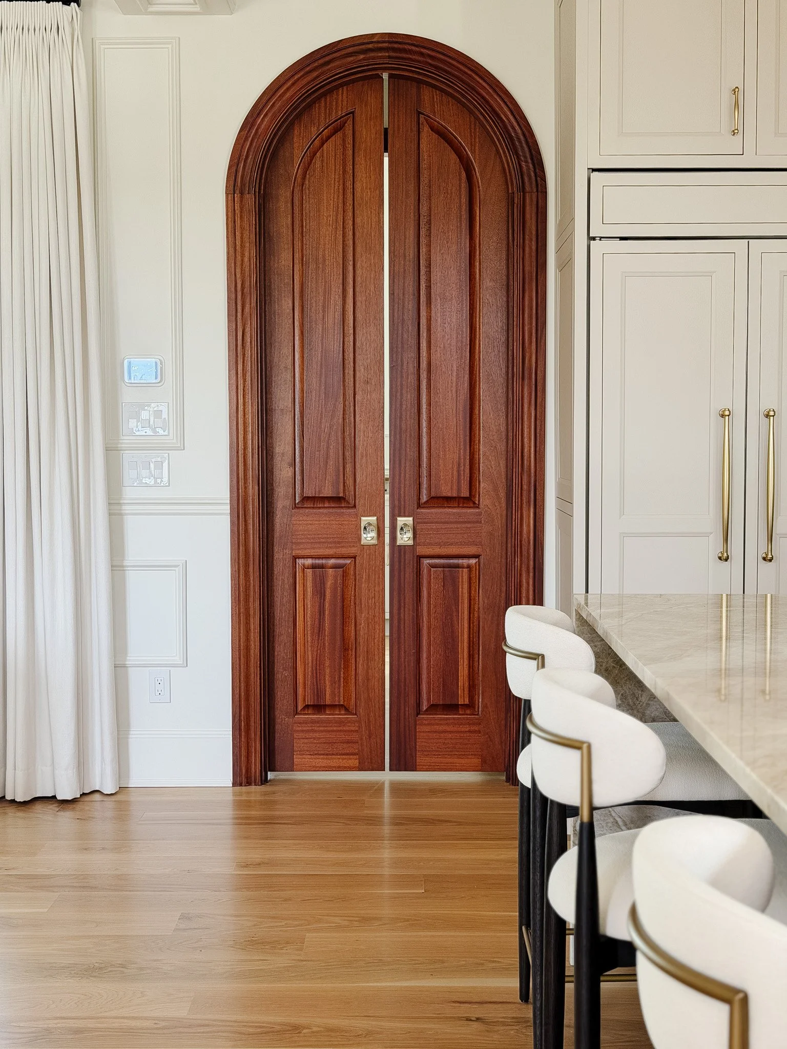 Interior view of a kitchen with a large wooden arched double door, white cabinetry with gold handles, and a marble island with three white chairs with black legs.