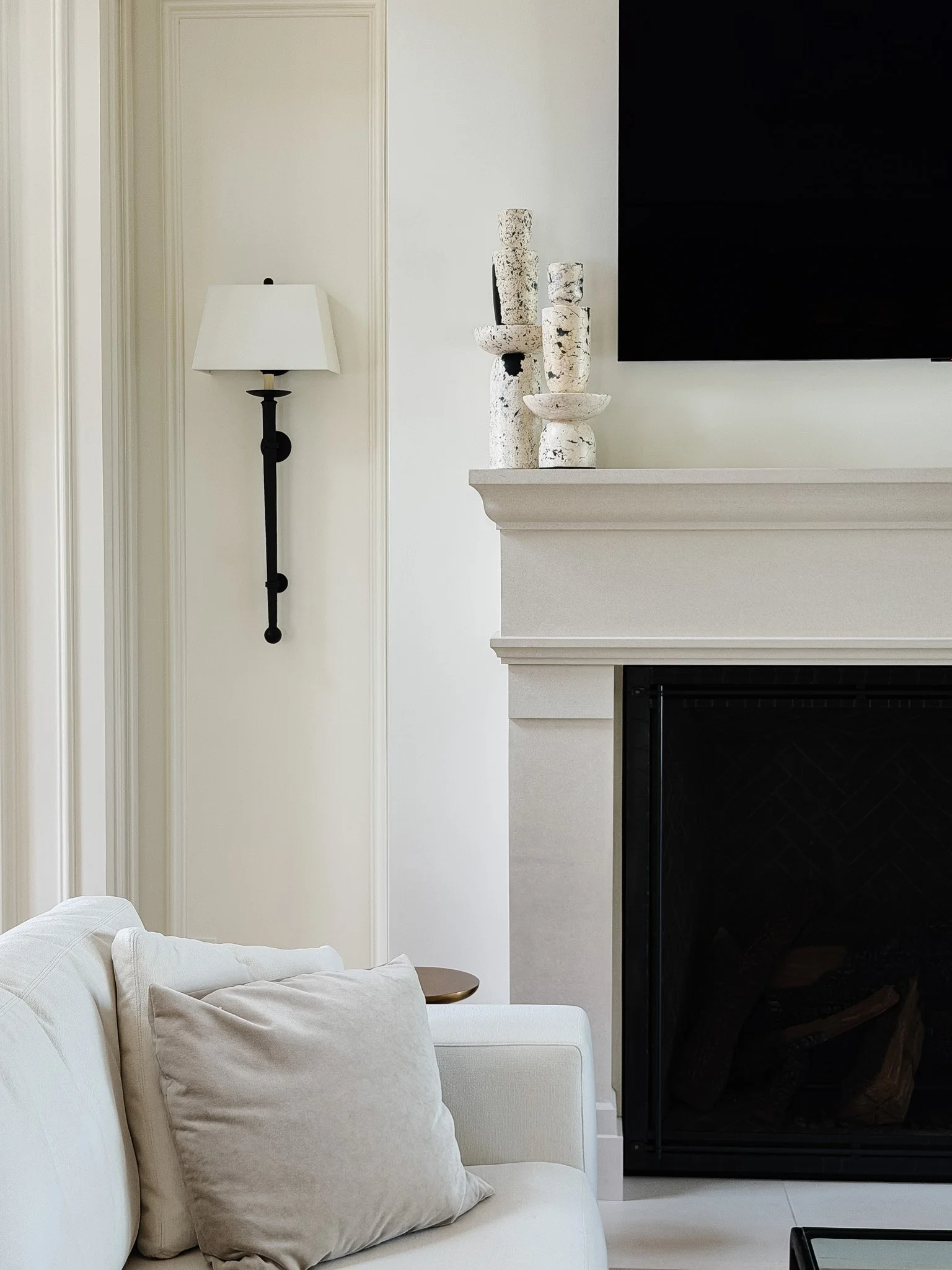 Living room with a cream sofa, beige cushions, a side table, a fireplace, a wall-mounted TV, and a modern black wall sconce with a white shade.