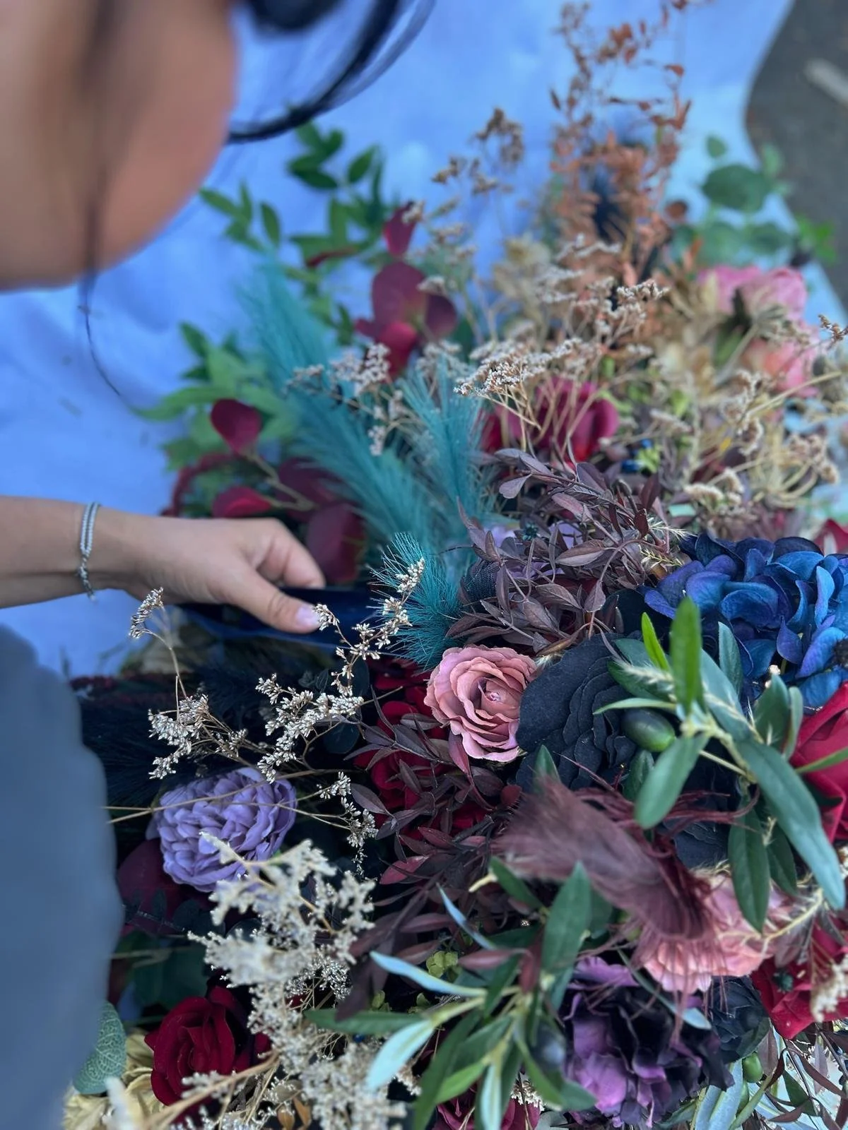 Person arranging a colorful bouquet of flowers including pink roses, purple hydrangeas, blue feathers, and assorted greenery on a table.