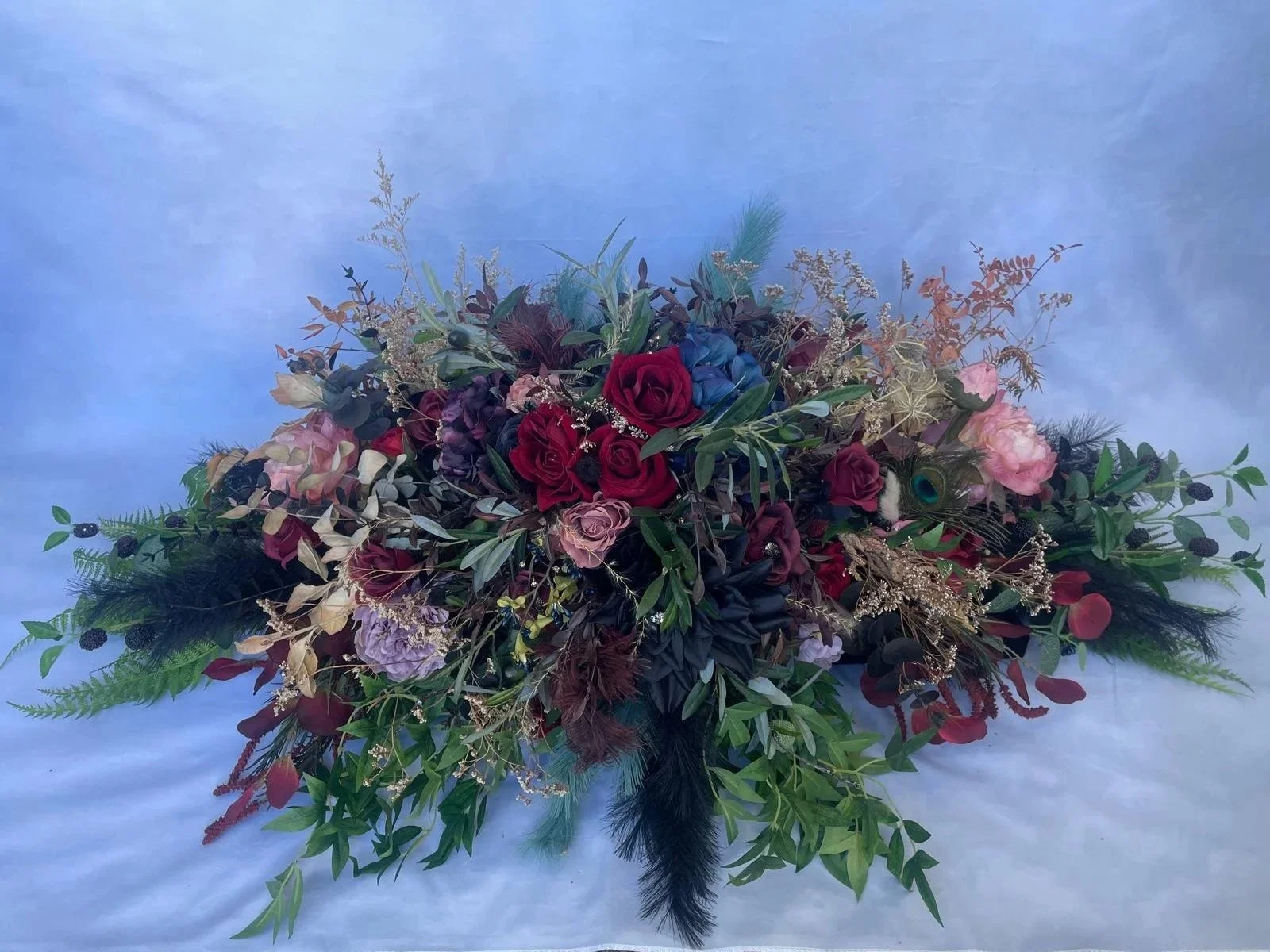 A floral arrangement with red roses, blue flowers, pink flowers, green leaves, and other foliage, displayed against a light blue background.