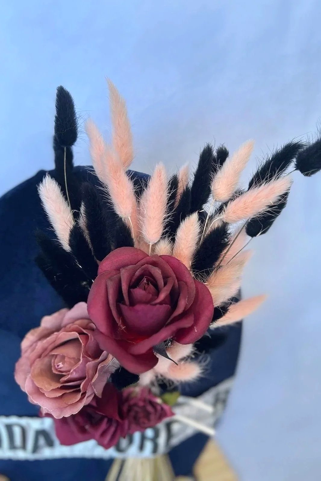 A bouquet of flowers with a large burgundy rose, a smaller pink rose, and various pink and black pampas grass against a light blue background.