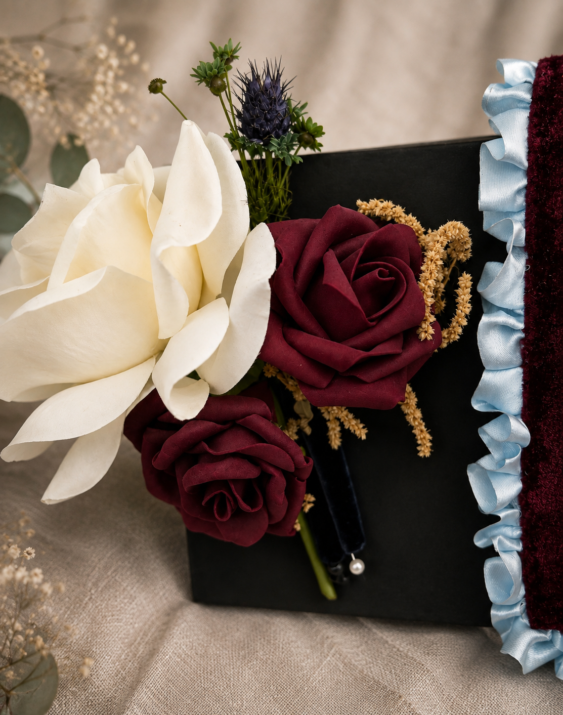 Close-up of a floral arrangement with white magnolia, red roses, small purple flower, and beige filler flowers in a black box with blue ribbon edging.