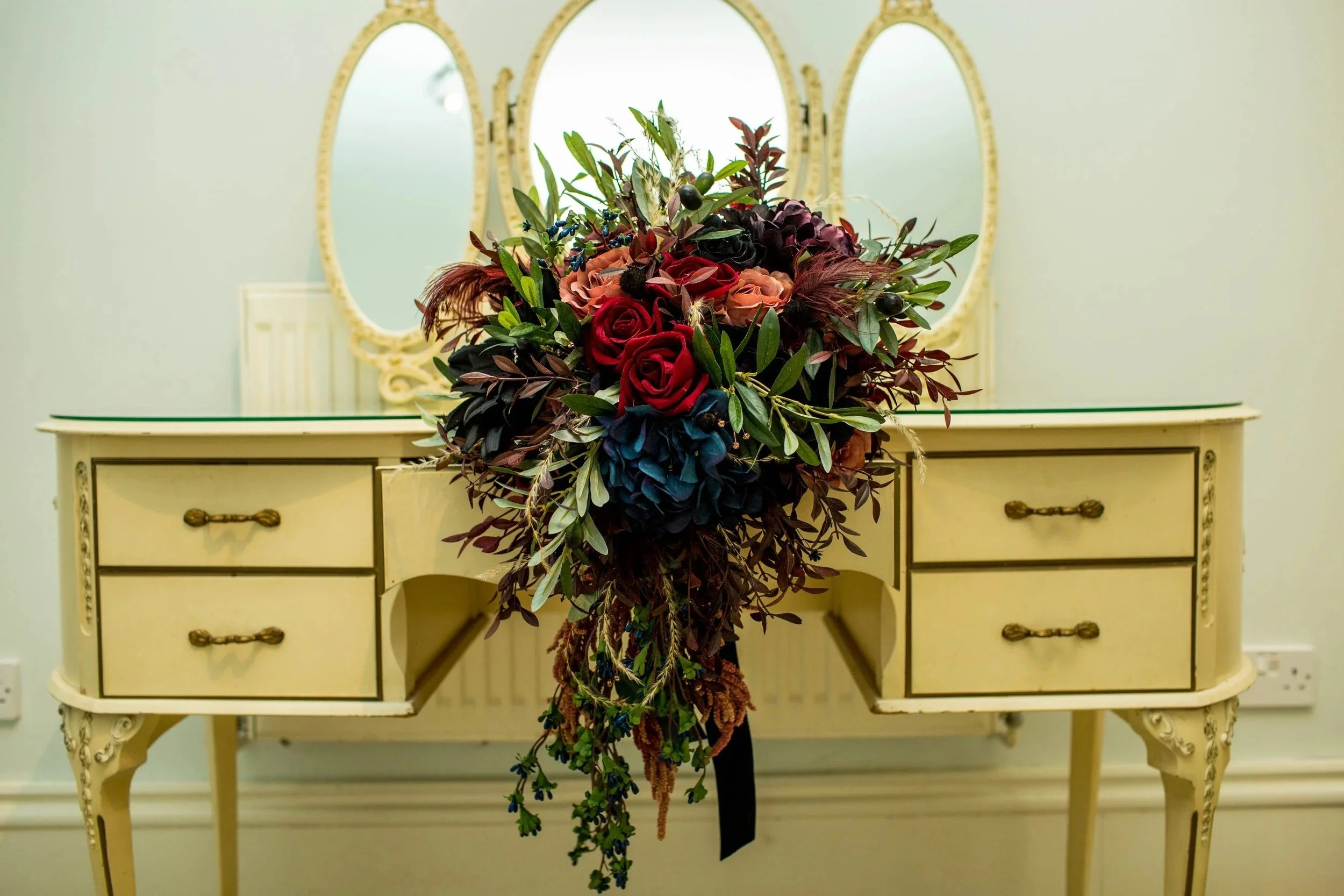 A bouquet of various flowers and greenery resting on a vintage cream-colored dresser with four drawers and ornate handles, placed against a light-colored wall with three mirrors above.