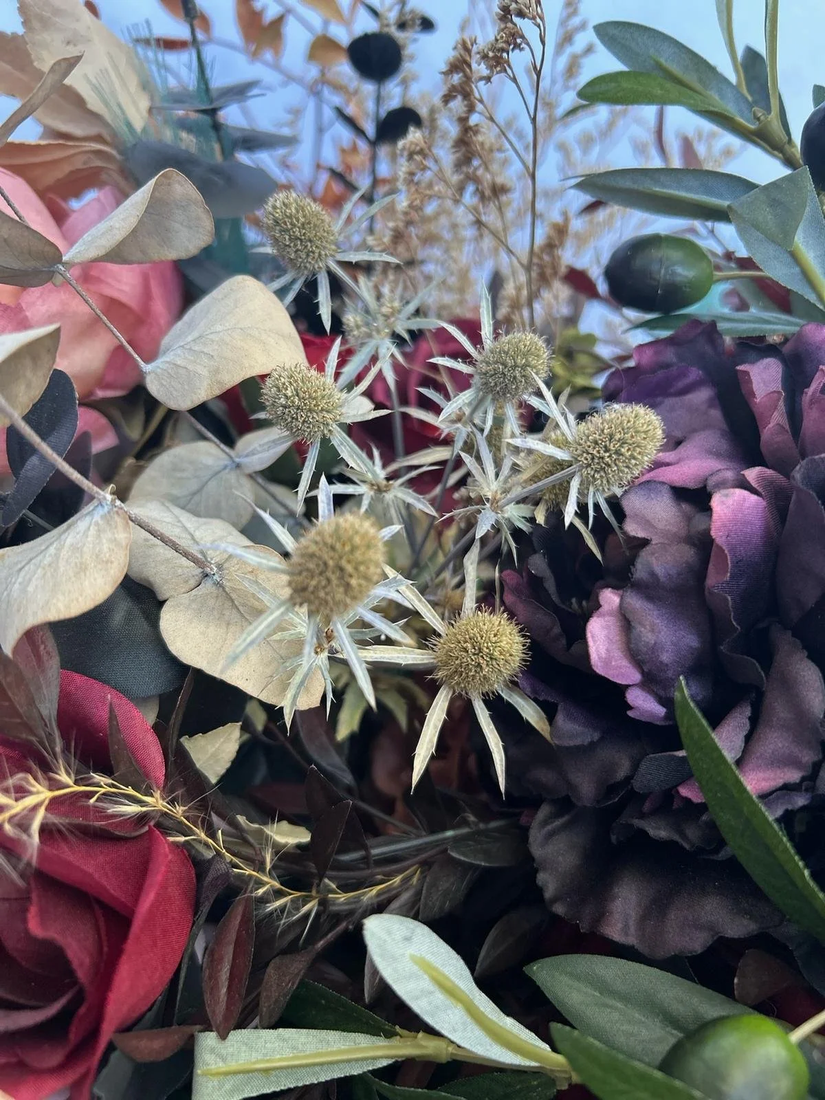 Close-up of a bouquet of dried and artificial flowers with green, purple, and beige foliage, featuring spiky seed pods and soft petals.