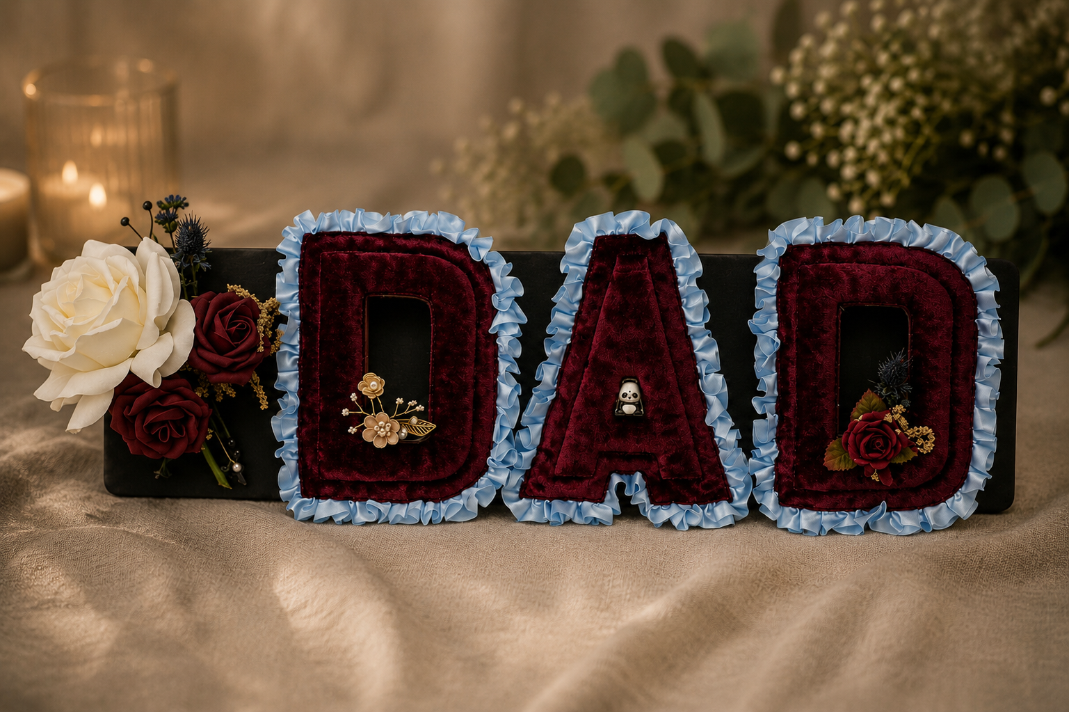 Decorative sign spelling 'DAD' with burgundy velvet letters, light blue ruffled borders, floral accents, and small panda and flower charms, placed on a beige fabric surface with candles and greenery in the background.