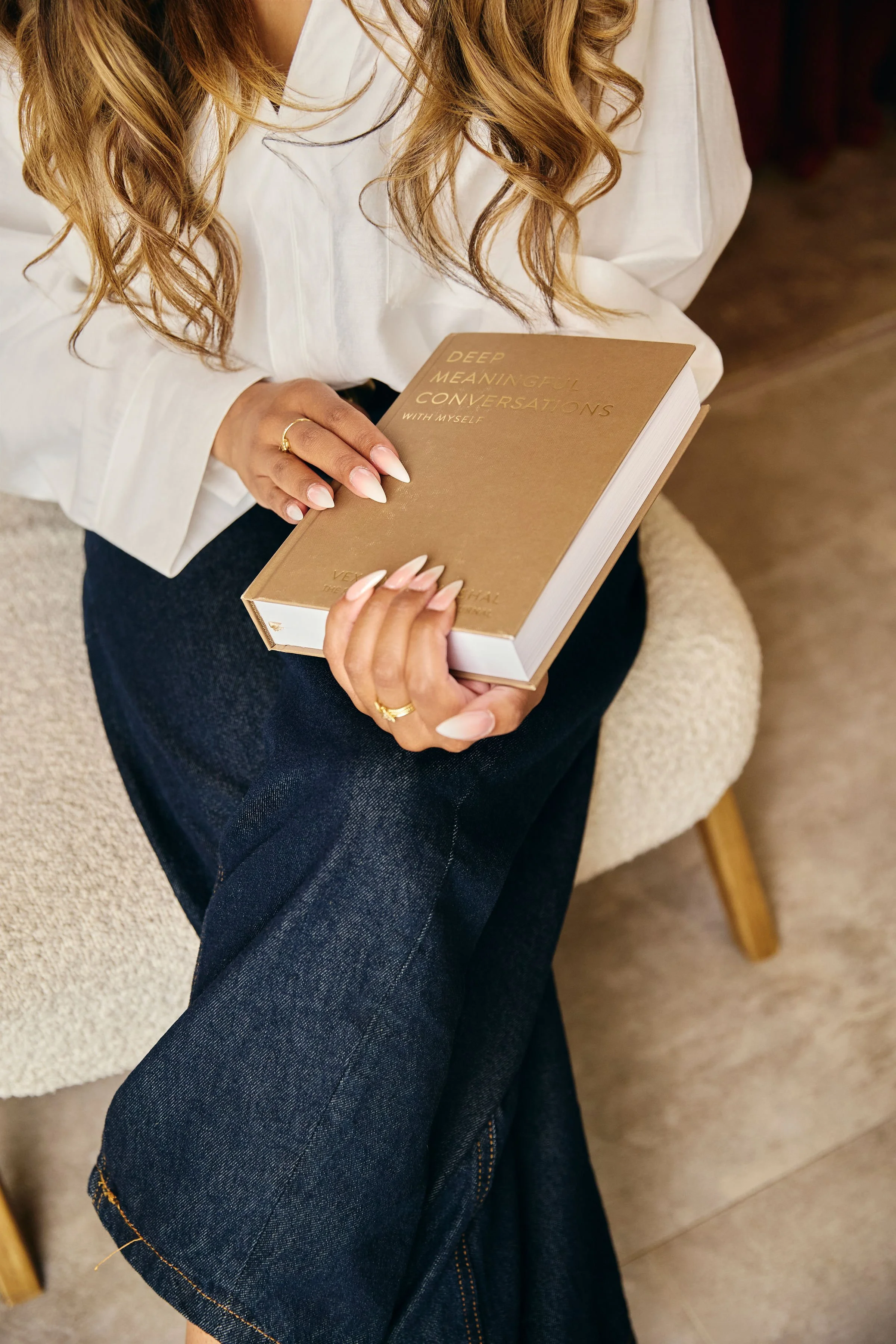 A woman with long wavy hair, wearing a white shirt and dark jeans, is sitting on a beige chair and holding a book titled 'Deep Meaningful Conversations with Myself.'