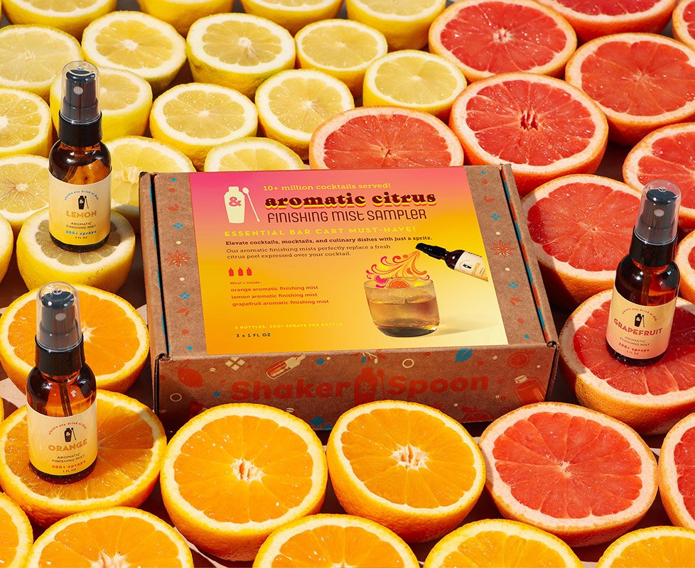 A box labeled 'Aromatic Citrus Finishing Mist Sampler' surrounded by halved lemons, oranges, and grapefruits. The box contains citrus-flavored finishing mists for cocktails.