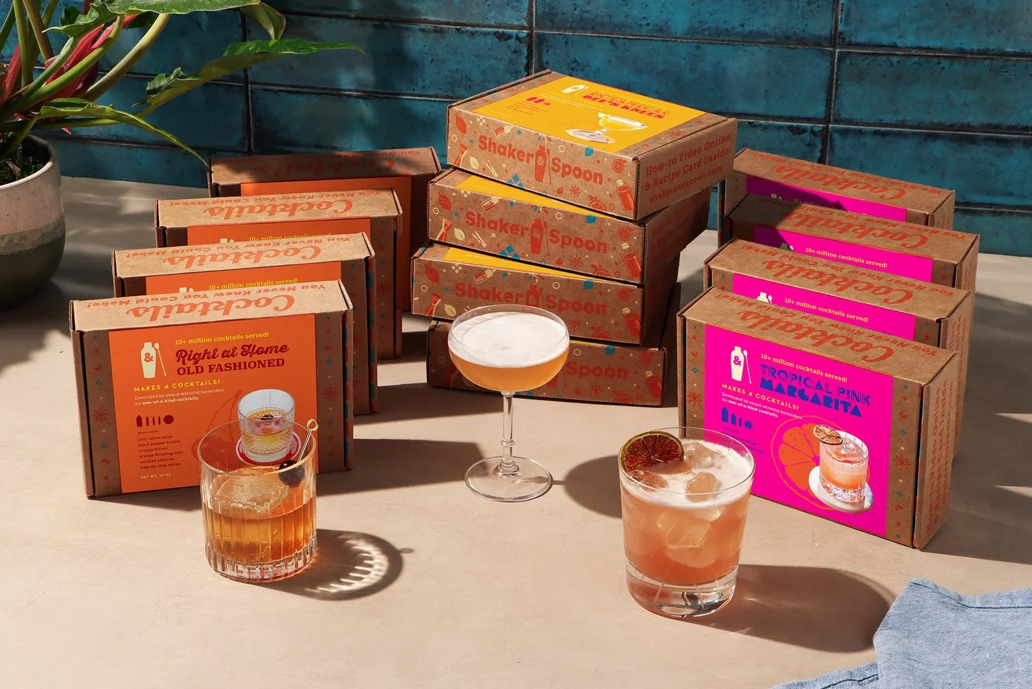 Multiple boxes of cocktail kits labeled 'Right at Home Old Fashione' and 'Tropical Pink Marietta' on a table, with two prepared cocktails in front, one with an orange slice and the other with a dried citrus garnish, a potted plant to the left, and a blue tiled wall in the background.