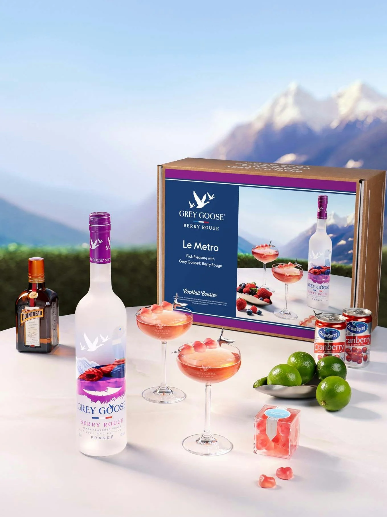 A display of Grey Goose Berry Rouge flavored vodka bottles, cocktail glasses with pink drinks, canned cranberry juice, fresh limes, and a box of Grey Goose Berry Rouge cocktail mix against a mountain landscape background.
