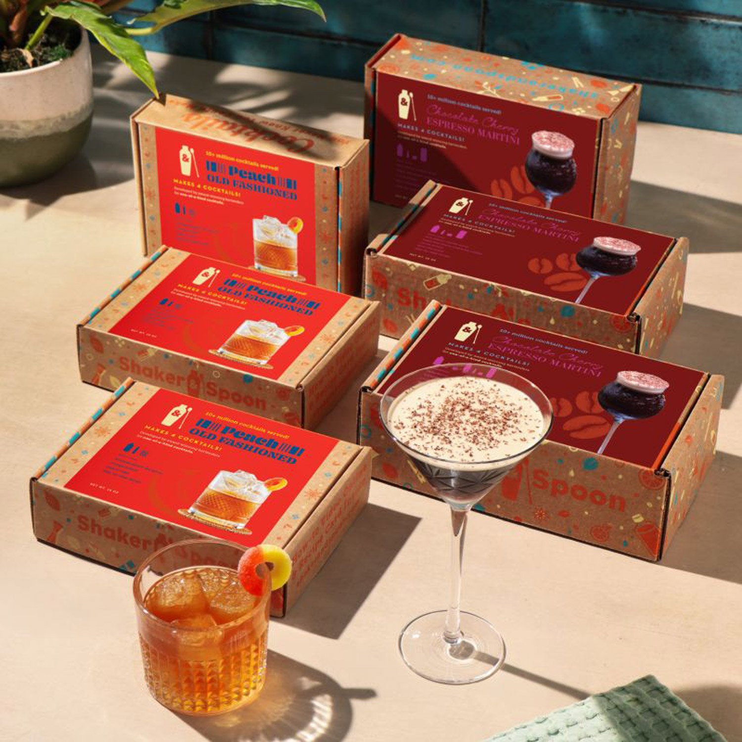 Multiple boxes of cocktail mixes on a table, along with a martini glass filled with a white creamy drink garnished with cocoa powder, and a glass of orange cocktail with a cherry garnish, in a well-lit outdoor setting with a potted plant nearby.