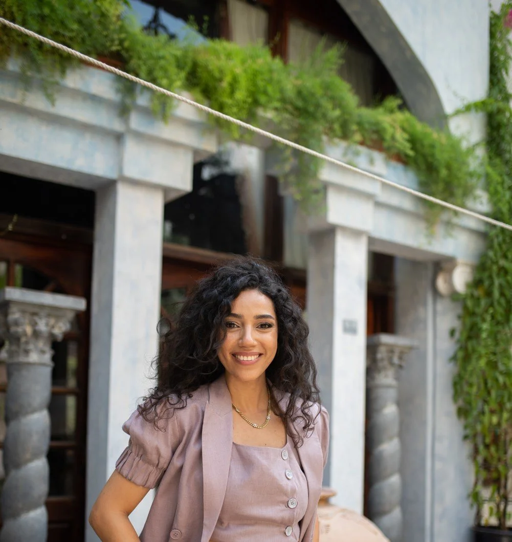 A woman with curly dark hair smiling outdoors in front of a building with columns and greenery.