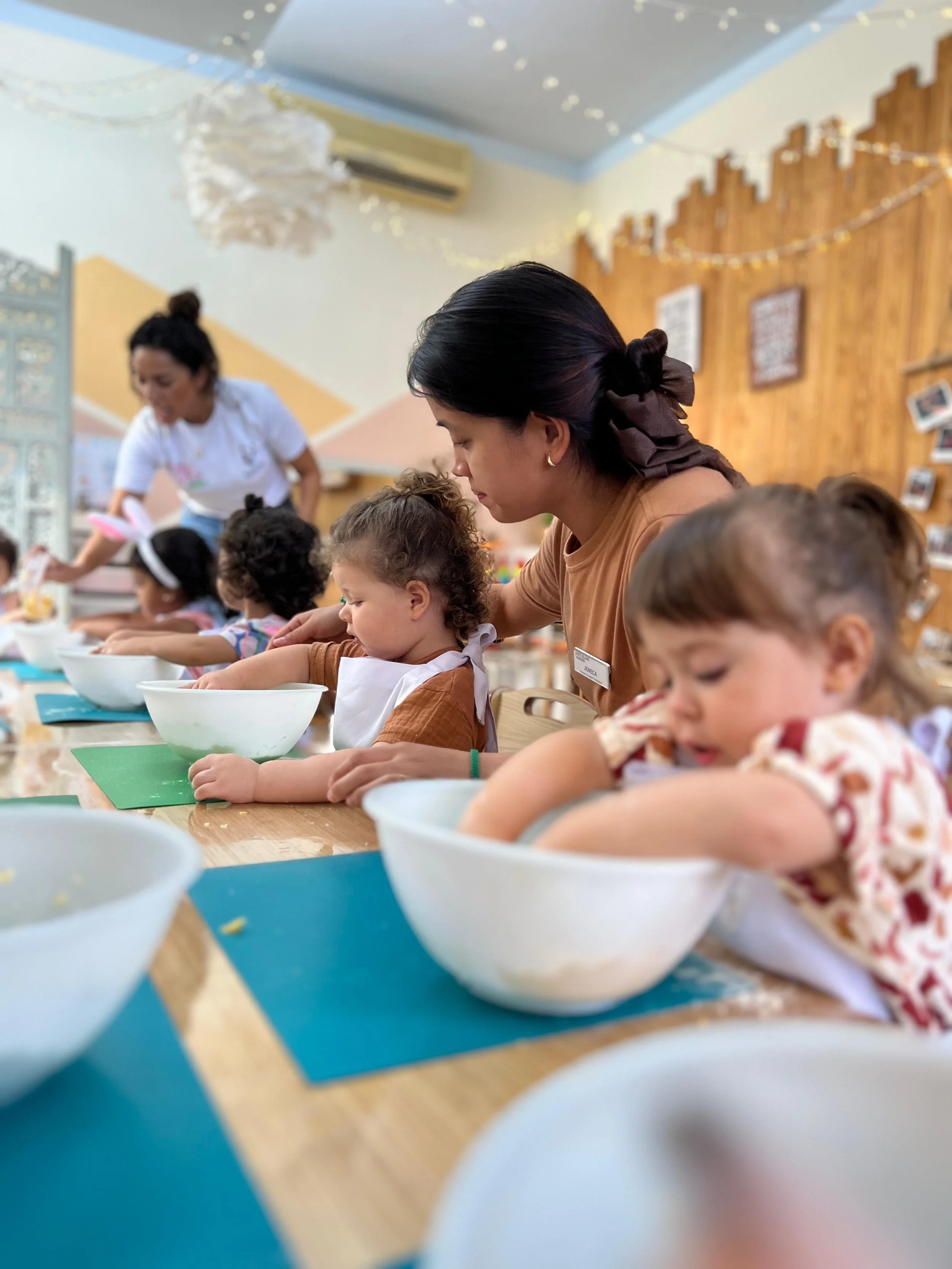 Children and adults participating in a cooking or baking activity at a communal table, with bowls, utensils, and paper mats visible.