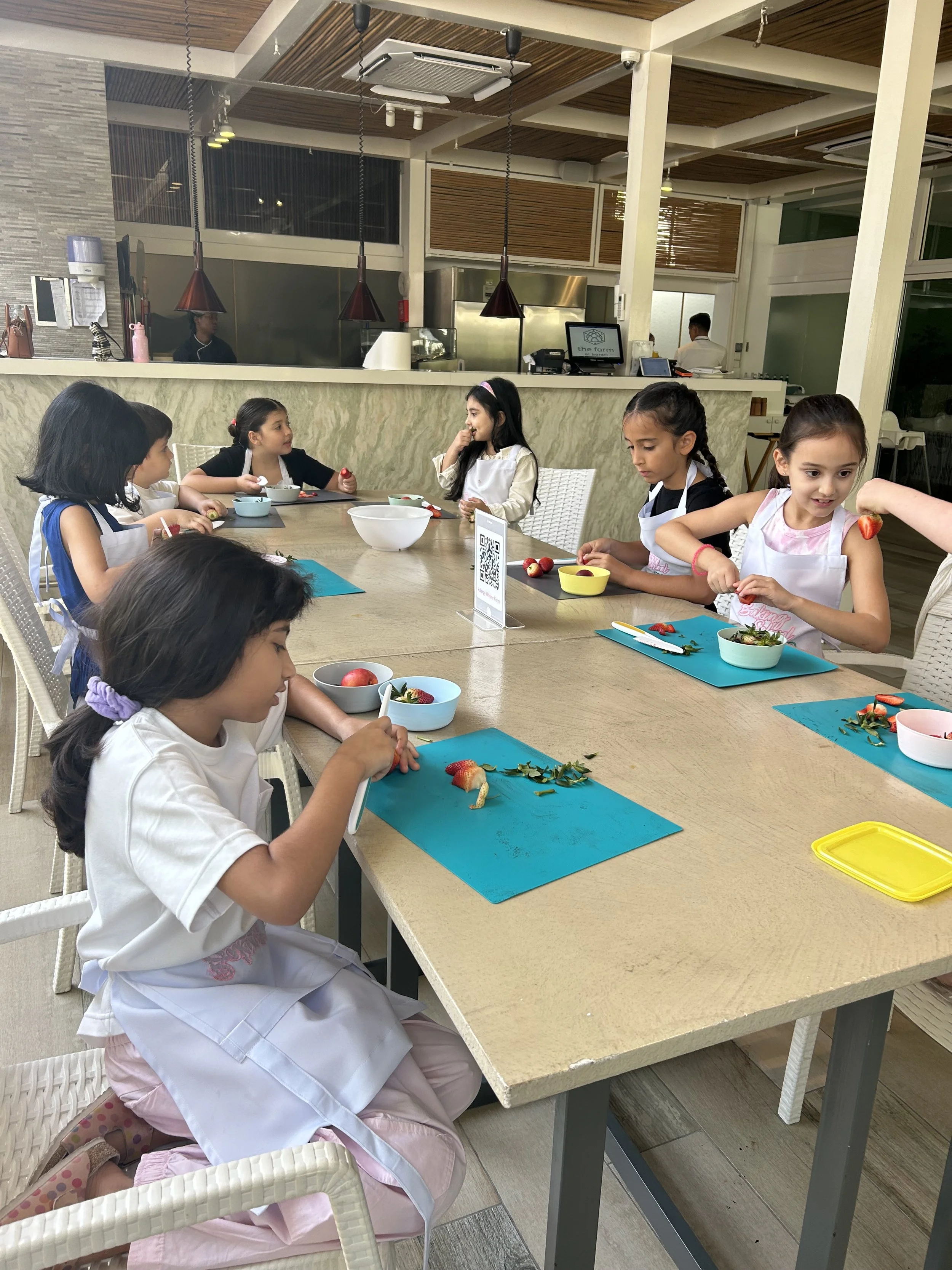 Children sitting at a long table in a restaurant, engaging in a cooking activity with strawberries and greenery on blue placemats, while a kitchen is visible in the background.