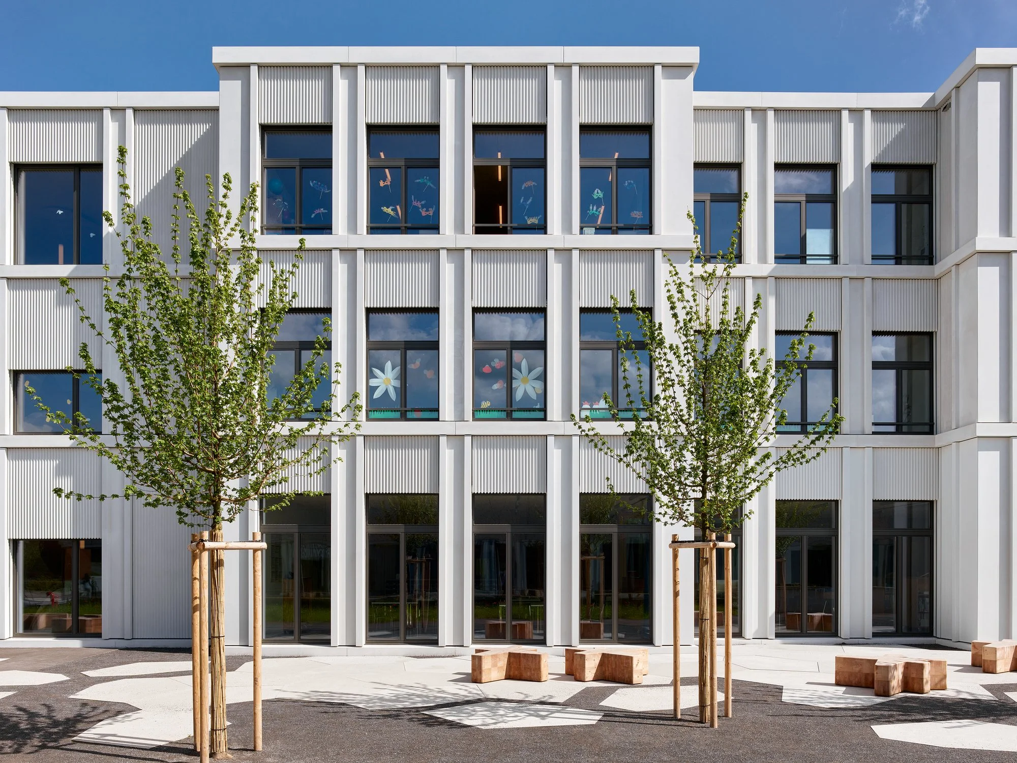 Modern white building with large windows decorated with colorful drawings; two young trees in front with wooden supports and some scattered wood blocks on the ground; clear blue sky.