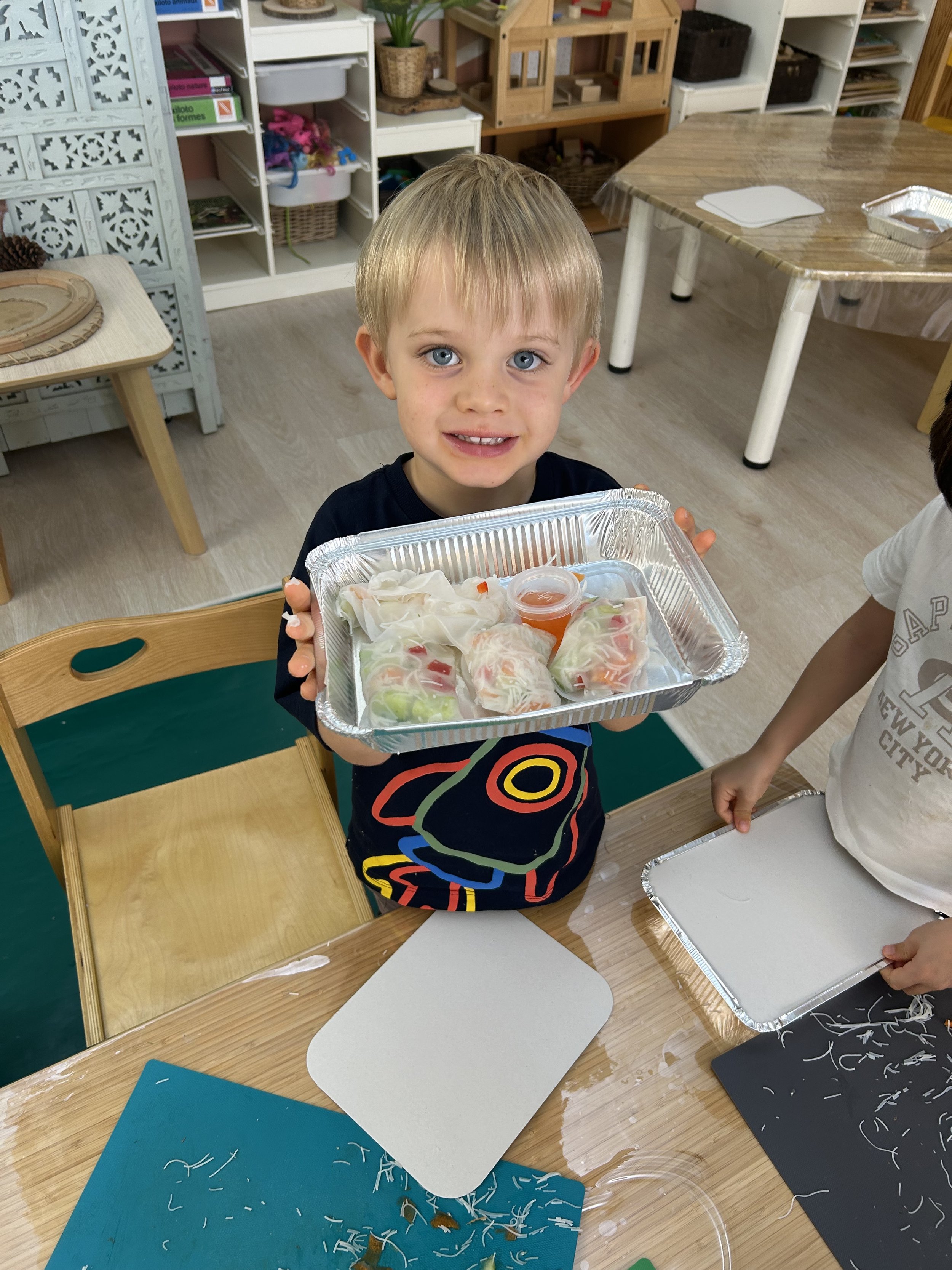 A young boy with blonde hair and blue eyes is holding a tray of spring rolls and a small container of sauce, smiling at the camera in a classroom or craft room.