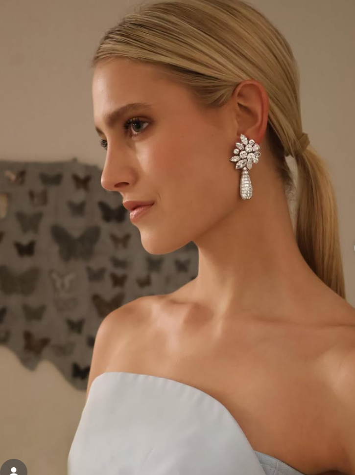 A woman with blonde hair styled in a low ponytail is wearing a pair of Adler diamond earrings and a strapless white top. The background features a wall decorated with butterfly silhouettes.