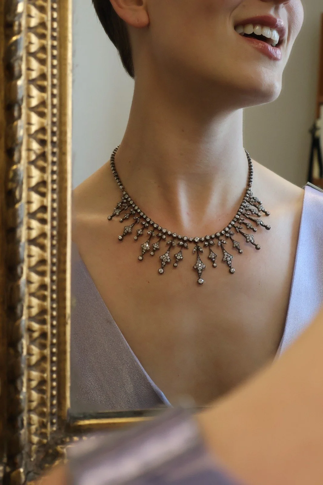 Close-up of a woman wearing a Victorian silver-topped gold sparkling diamond necklace, reflected in an ornate gold mirror, smiling with parted lips, wearing a satin lavender dress.