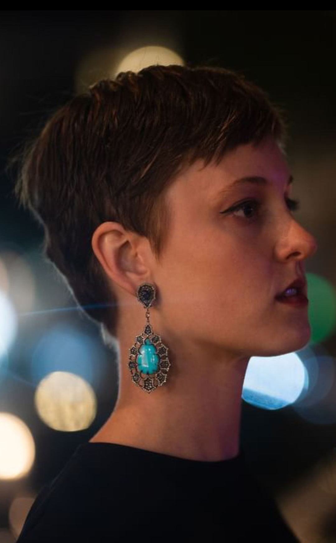 Profile of a woman with short brown hair, wearing a large turquoise and black ornate earring, in a nighttime setting with blurred city lights in the background.