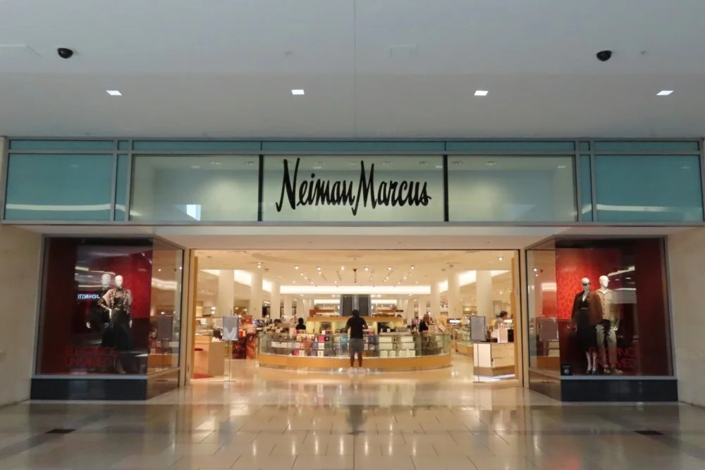 Neiman Marcus store entrance with mannequins in the display windows and interior shoppers visible inside.