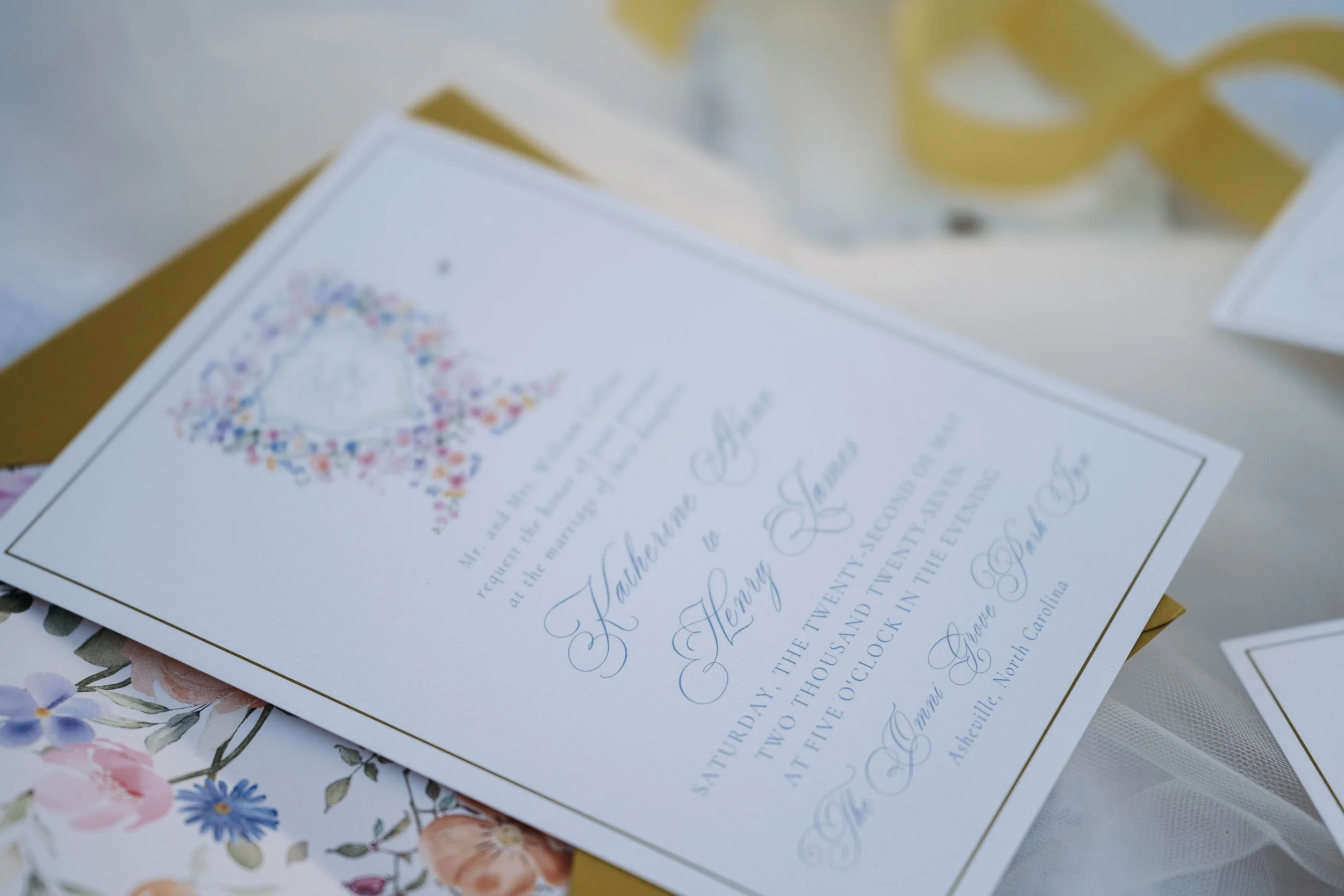 Close-up of a wedding invitation with elegant cursive writing and floral designs, partially visible on a white surface with a yellow object and other paper underneath.