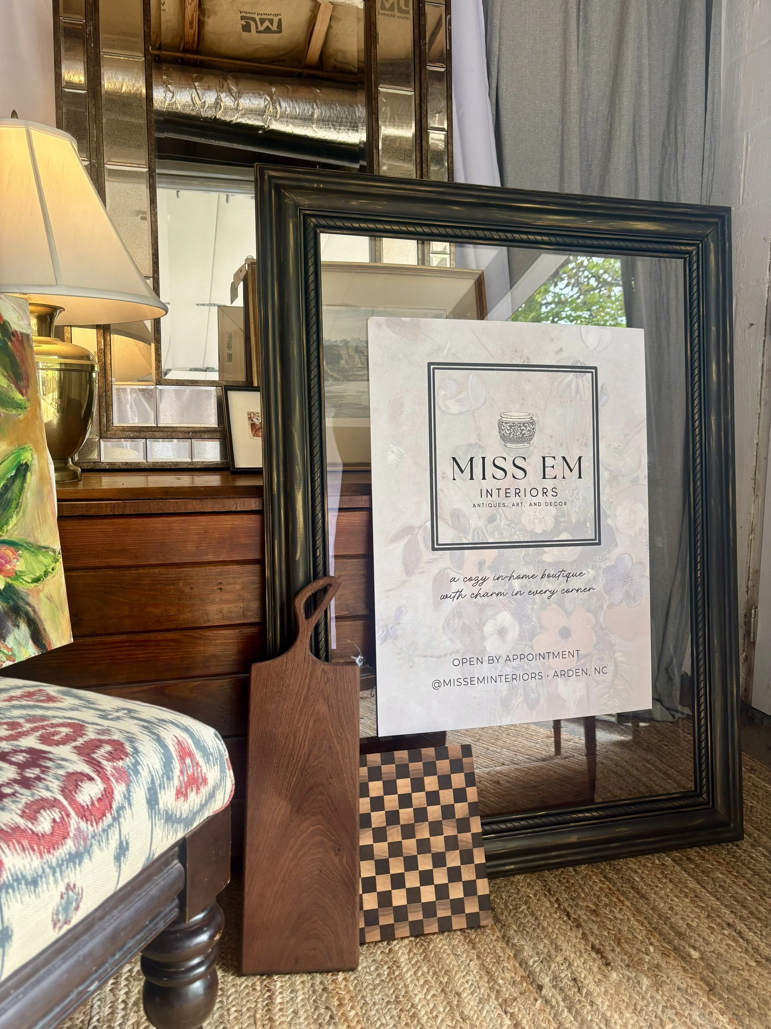 Interior decor items including a floral-patterned upholstered armchair, a table lamp, picture frames, a mirror, and a sign for Miss Em Interiors on a stand, with wooden furniture and a rug in the background.