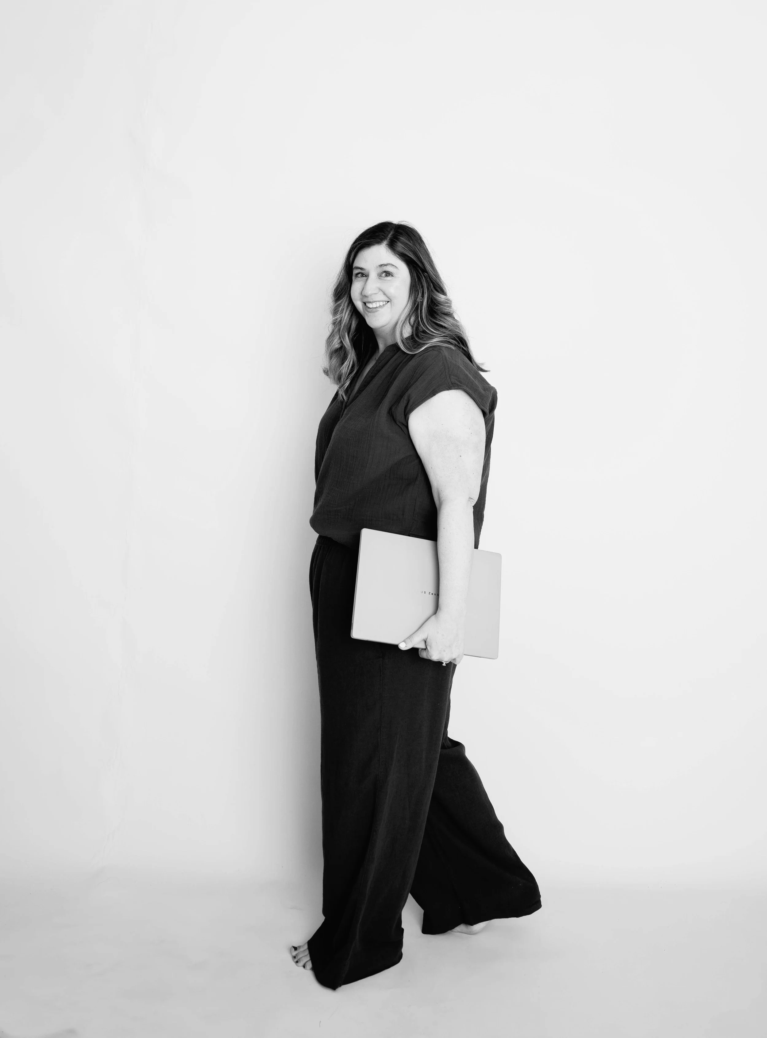 Black and white portrait of a woman with wavy hair, smiling, wearing a dark top and wide-legged pants, holding a laptop under her arm, standing against a plain white background.