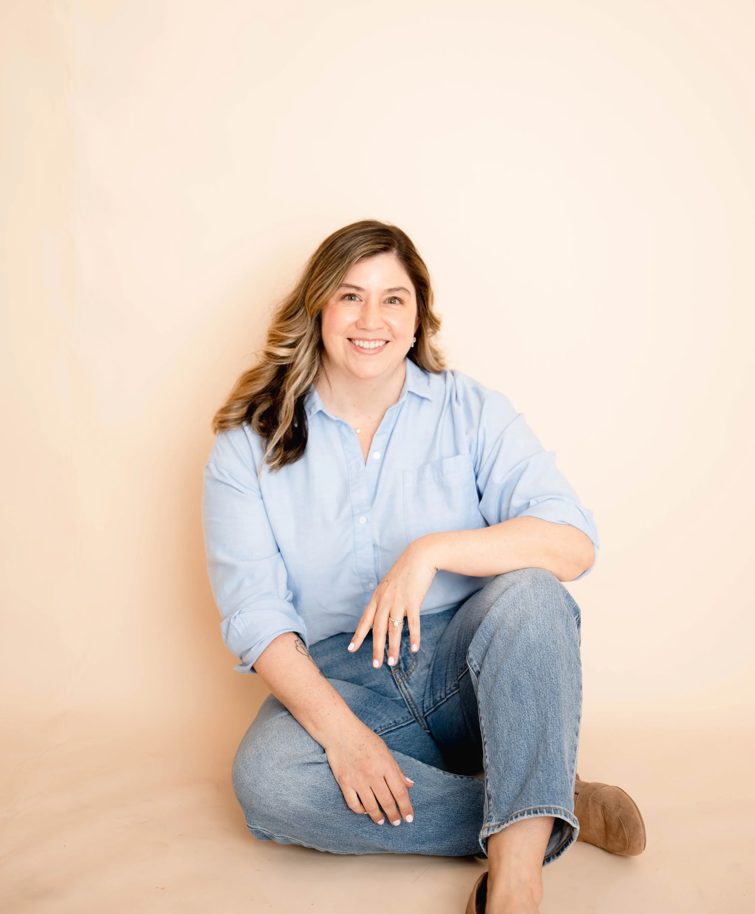 A woman with long wavy hair, wearing a light blue shirt and blue jeans, smiling while sitting on a beige background with her legs crossed.