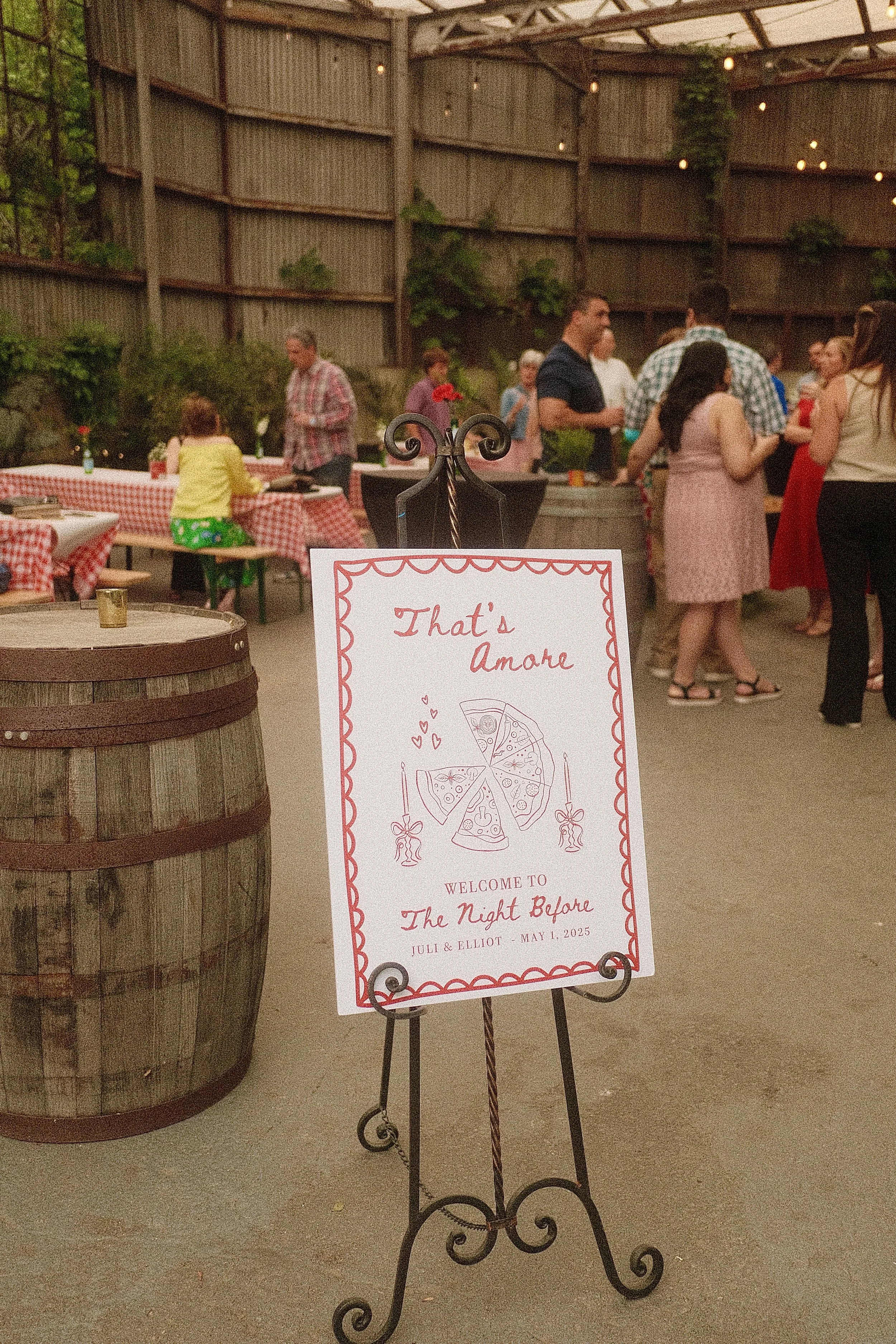 A sign for an event called "That's Amore" at The Night Before, with a pizza illustration, on an easel, in front of a party with people socializing under string lights in a rustic outdoor setting.