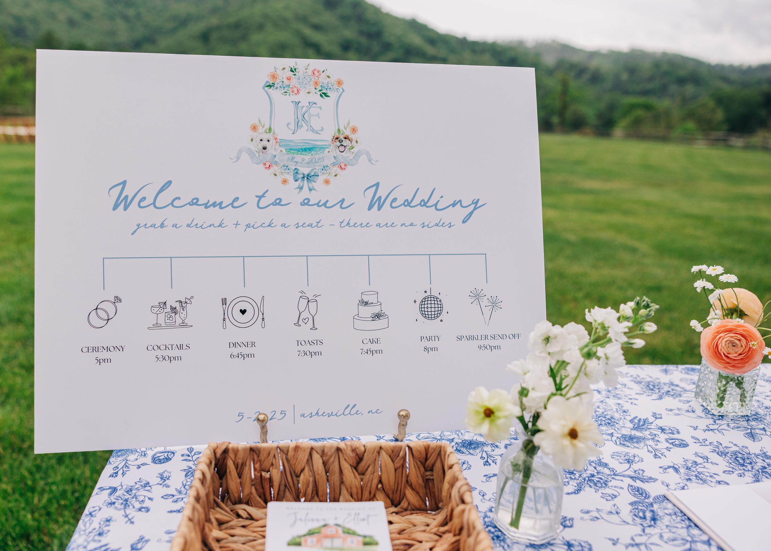 Wedding event signage with floral design, monogram, and event schedule in outdoor setting with flowers and greenery.