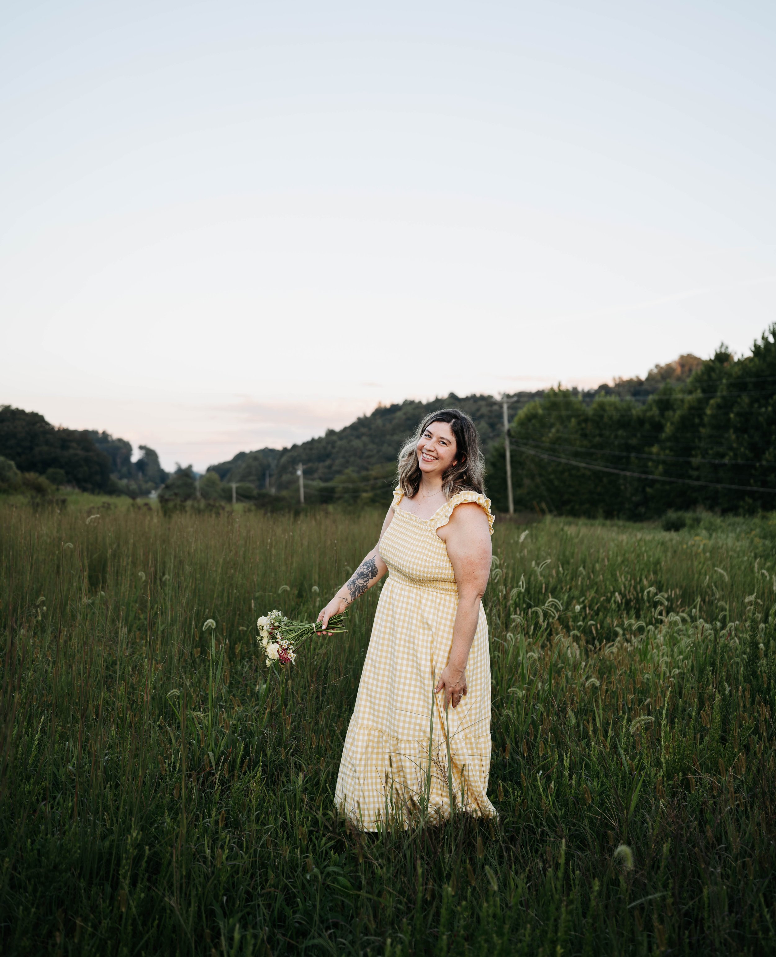 A woman standing in a grassy field holding a bouquet of flowers, with a backdrop of trees, hills, and a sky during daytime.