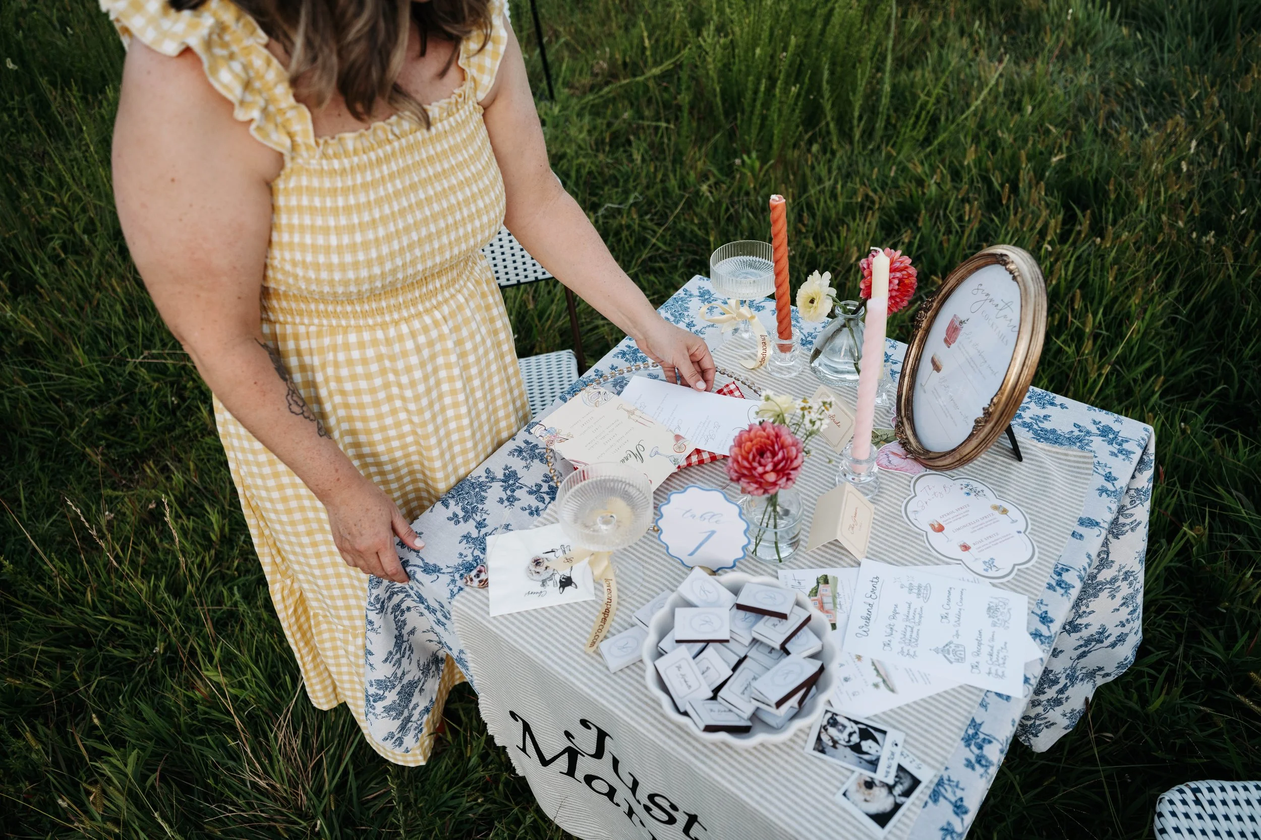 A woman in a yellow gingham dress organizing a wedding reception table outdoors, decorated with floral arrangements, candles, and printed wedding info.