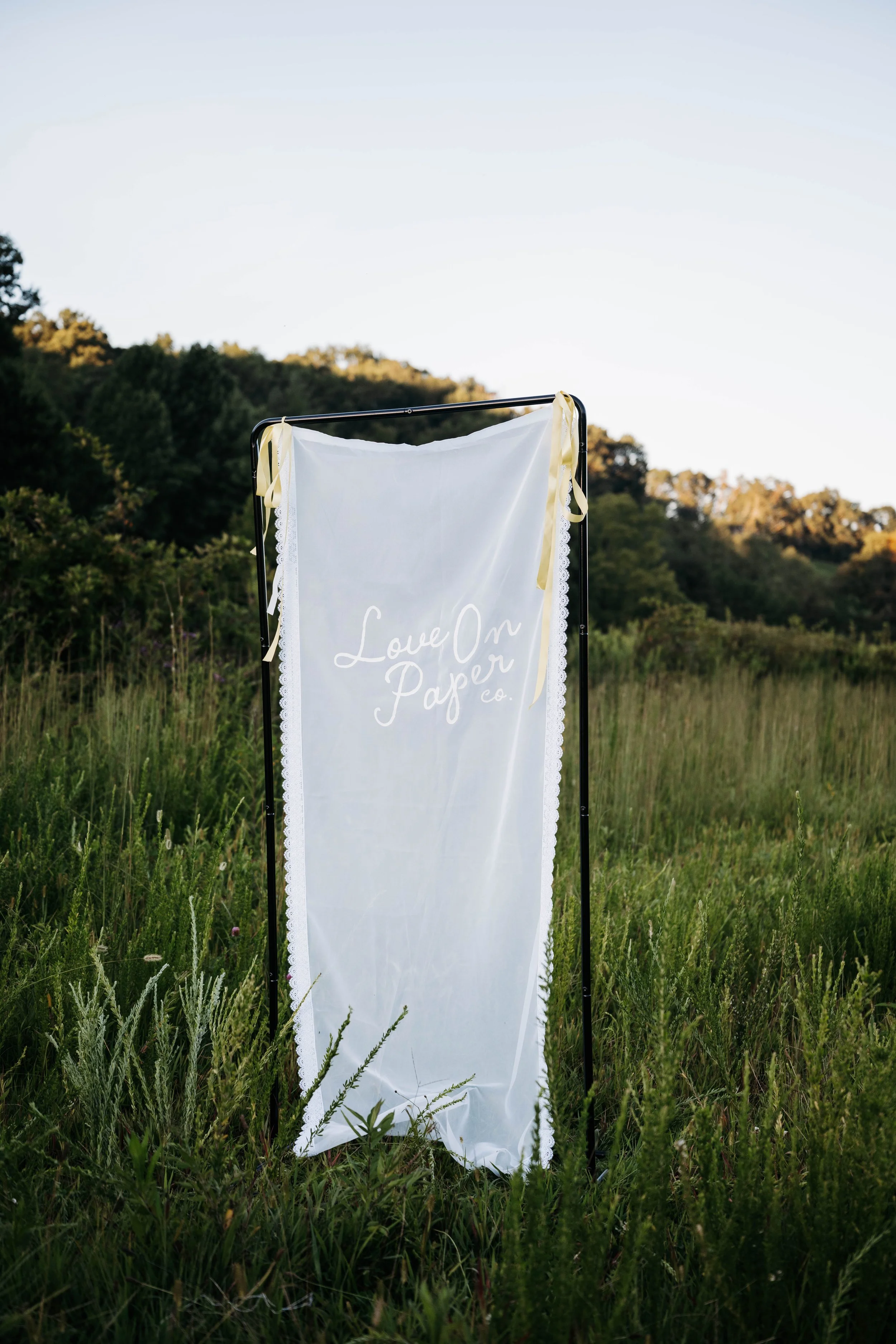 A white fabric banner with lace edges and yellow ribbons, displaying the text "Love on Paper Co." in a calligraphy style, hangs outdoors in a grassy field with trees and hills in the background.