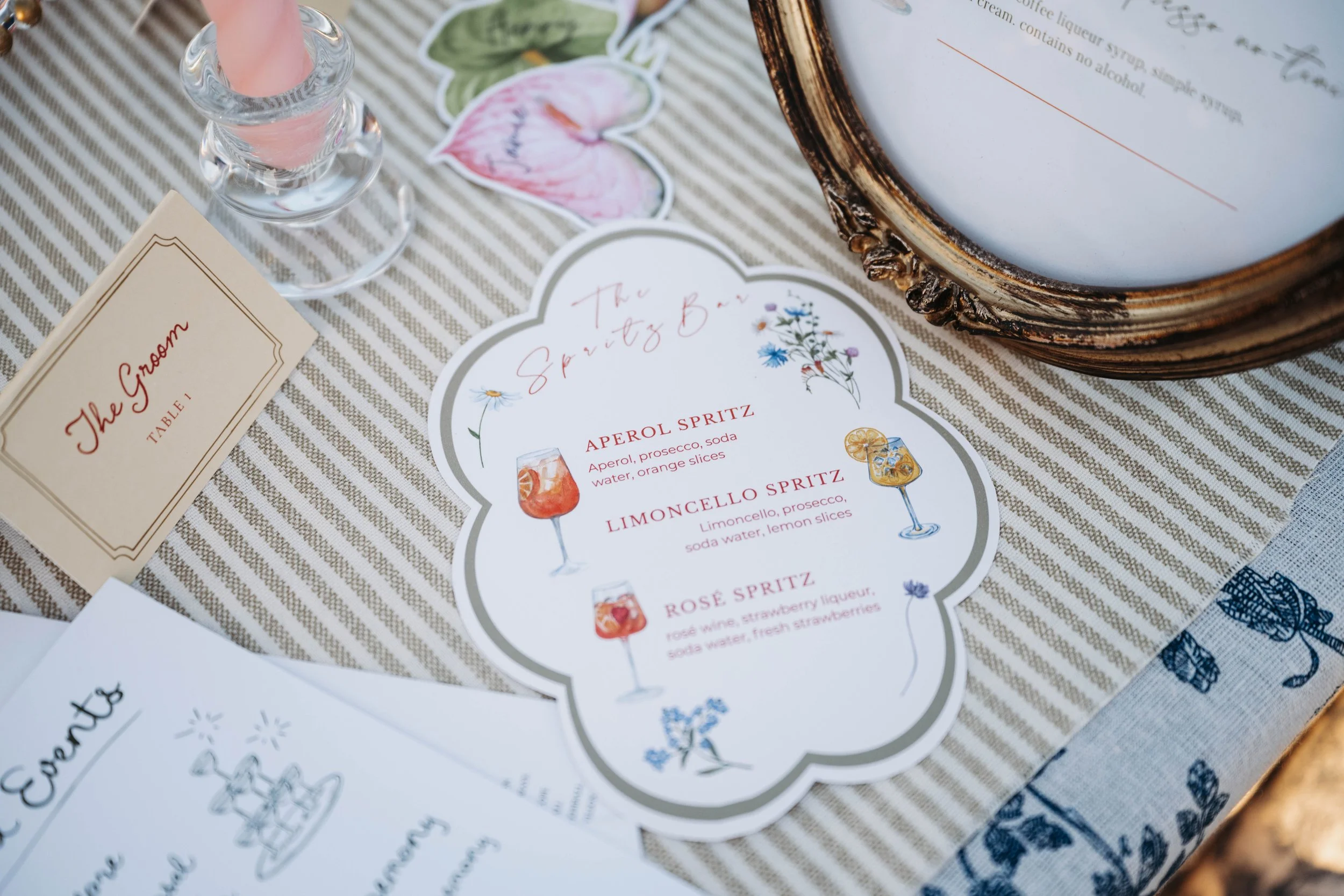 Close-up of a table with a floral-themed menu featuring various spritz cocktails, a small card labeled 'The Groom,' a glass of pink drink, and decorative paper cutouts of flowers and leaves on a striped tablecloth.