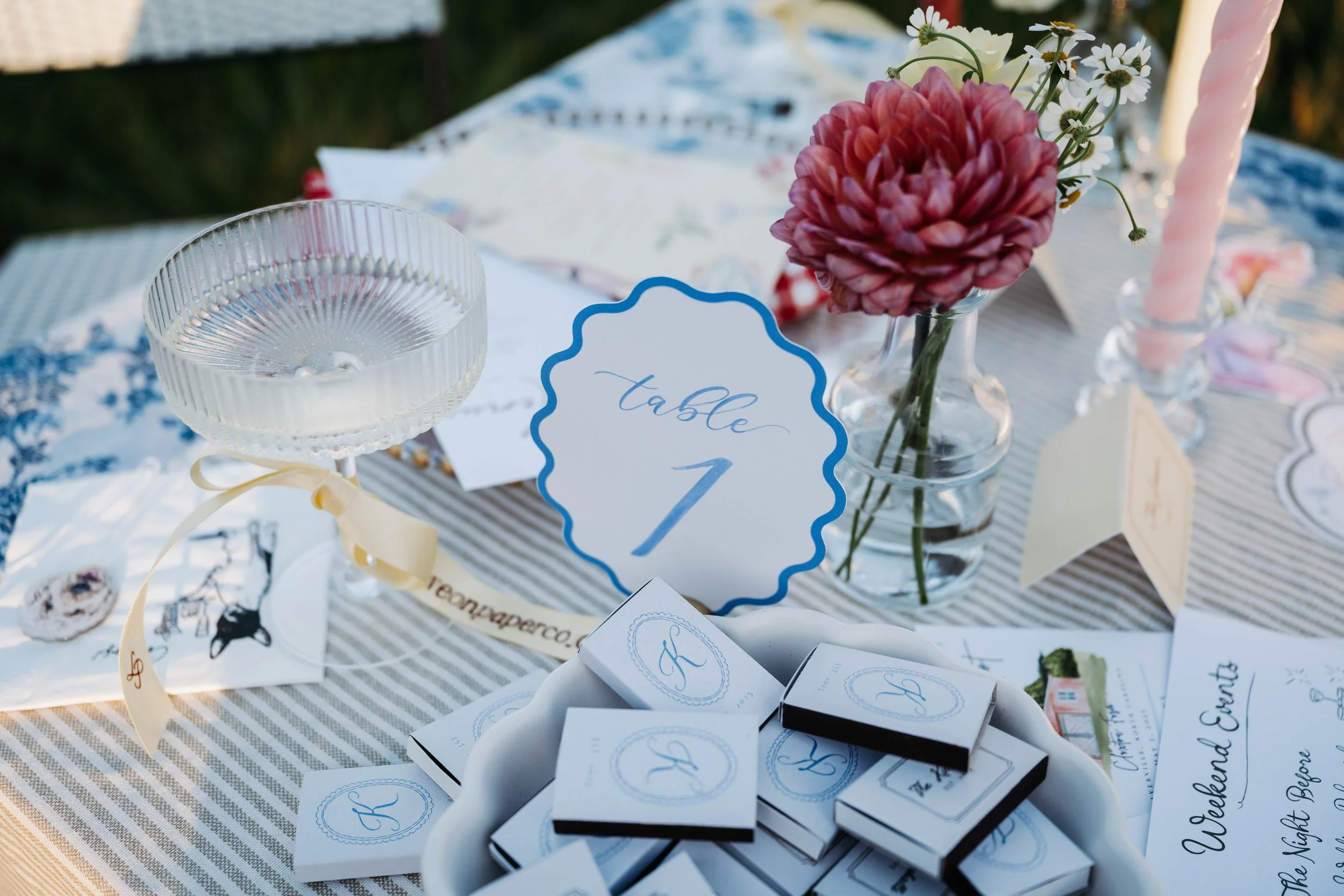 A table set outdoors with a floral centerpiece, including a pink dahlia and white daisies, surrounded by paper and boxed wedding favors with initials 'R' and elegant script messages.