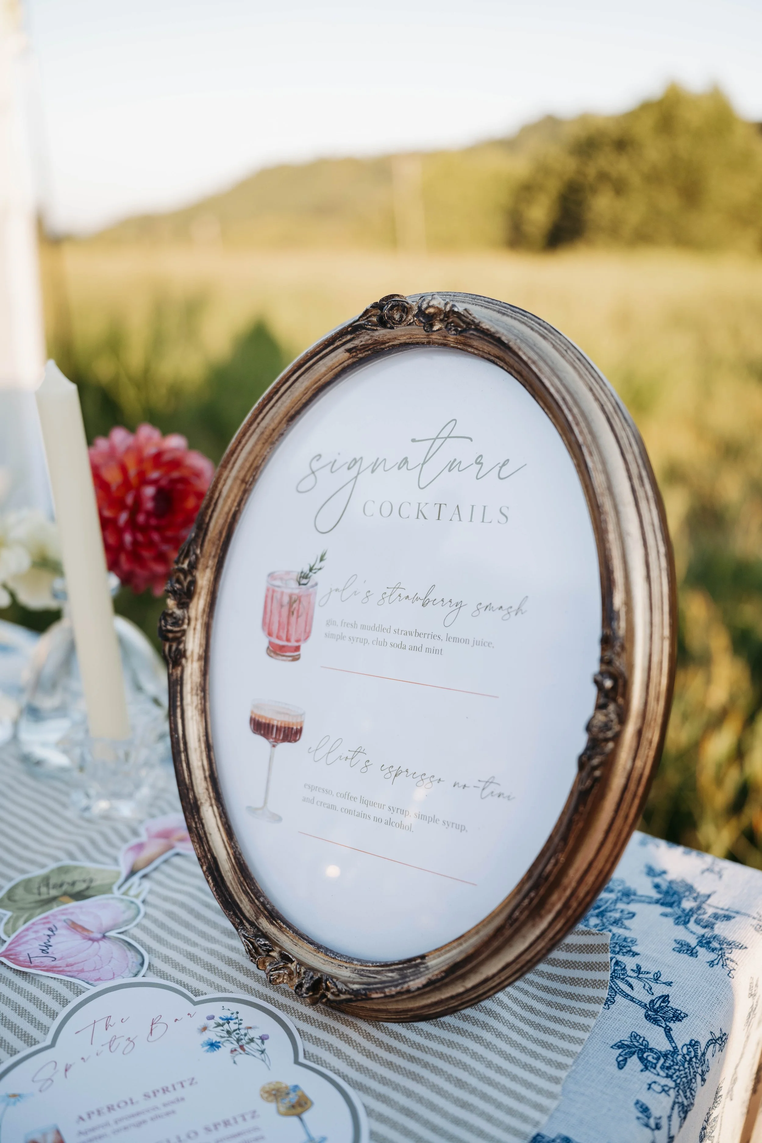 Vintage-style ornate picture frame holding a sign that reads 'Signature Cocktails' and lists drink recipes, placed on a striped tablecloth with floral accents and some floral decorations in the background.