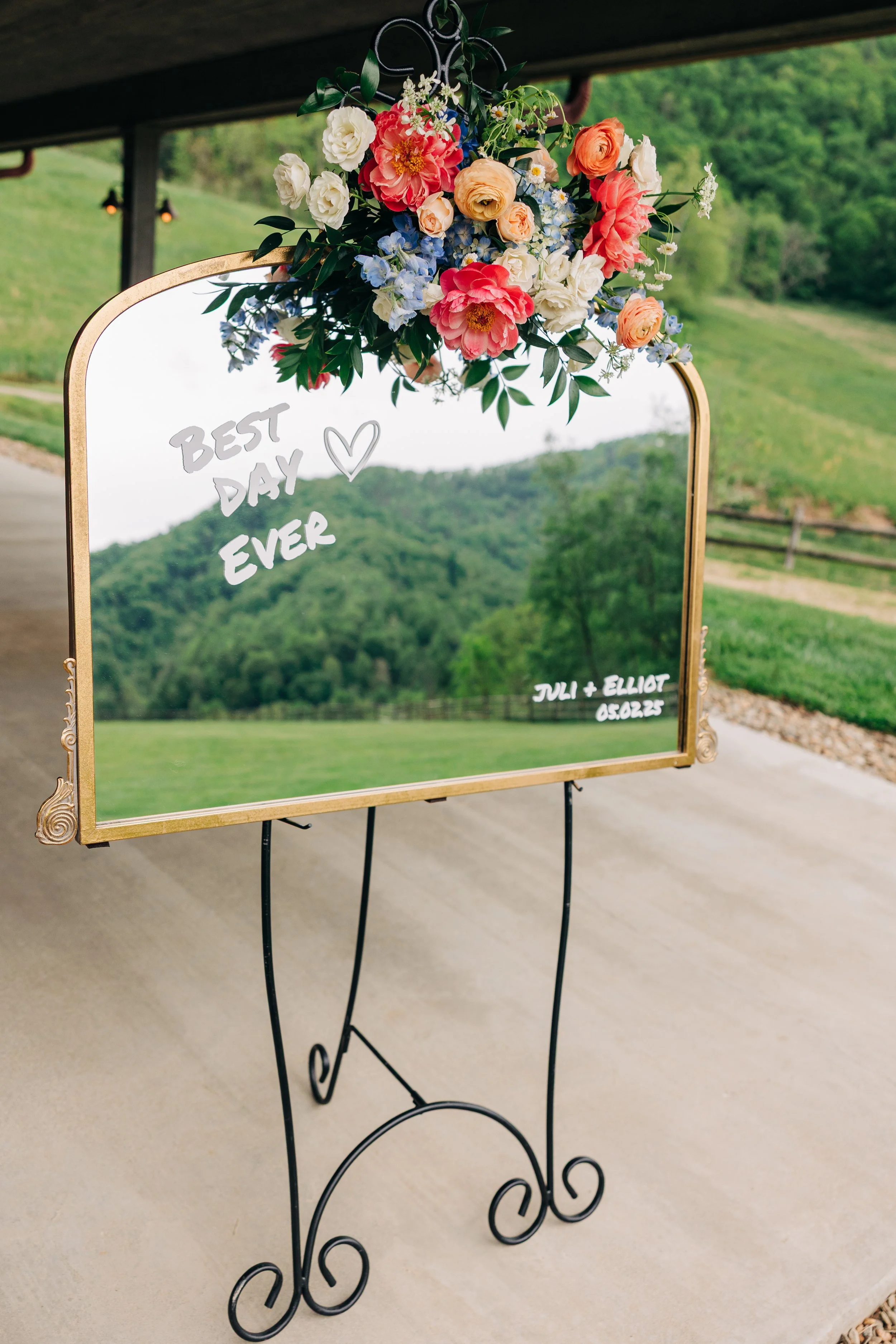 A decorative mirror with a gold frame and a floral arrangement on top, reflecting a scenic landscape of green hills and trees. Handwritten text on the mirror reads "Best Day Ever" with a heart, and "Juli + Elliot 05.02.25" in the bottom right corner.