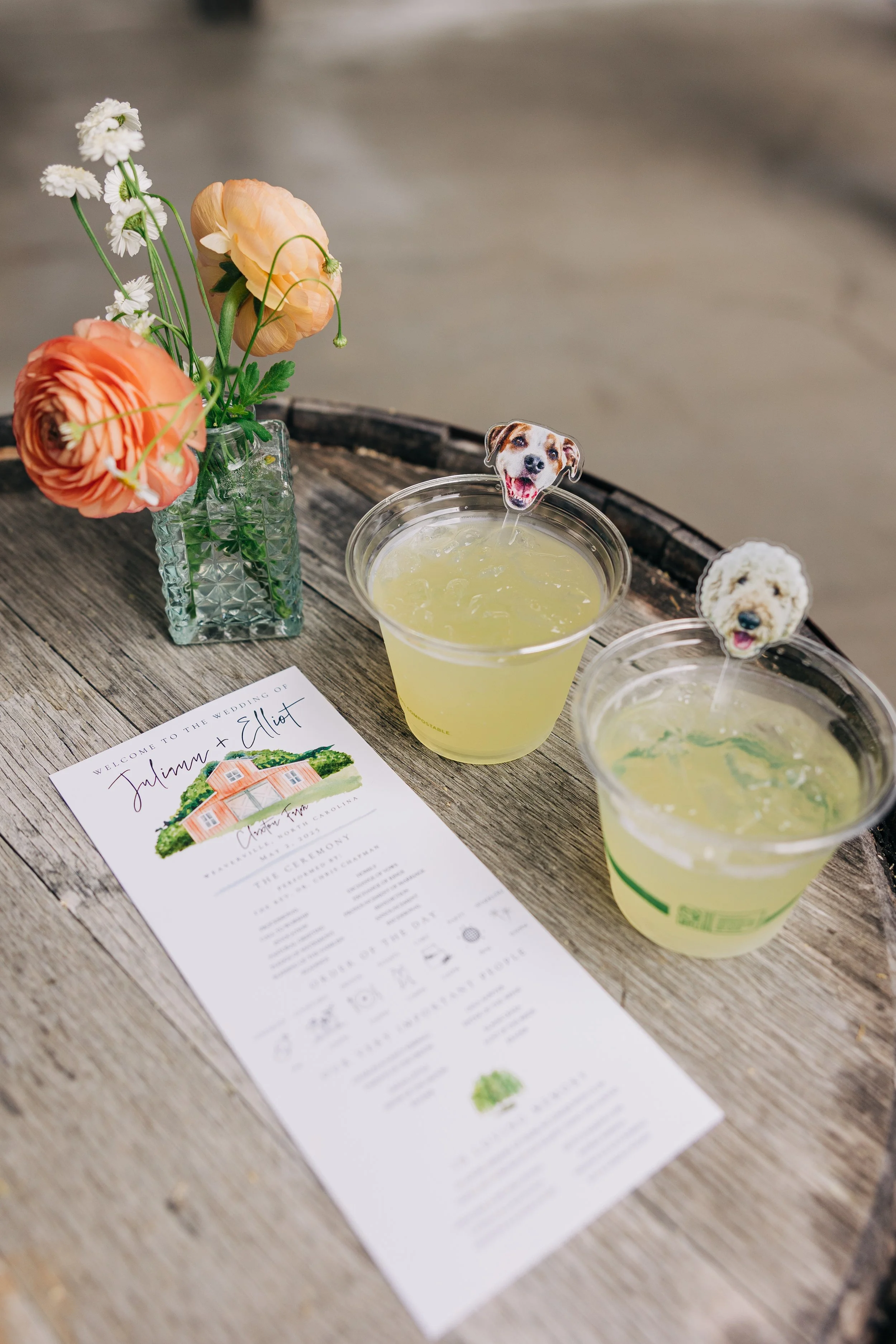 A bouquet of flowers in a textured glass vase, two drinks with dog-themed toppers, and a wedding program on a wooden table.