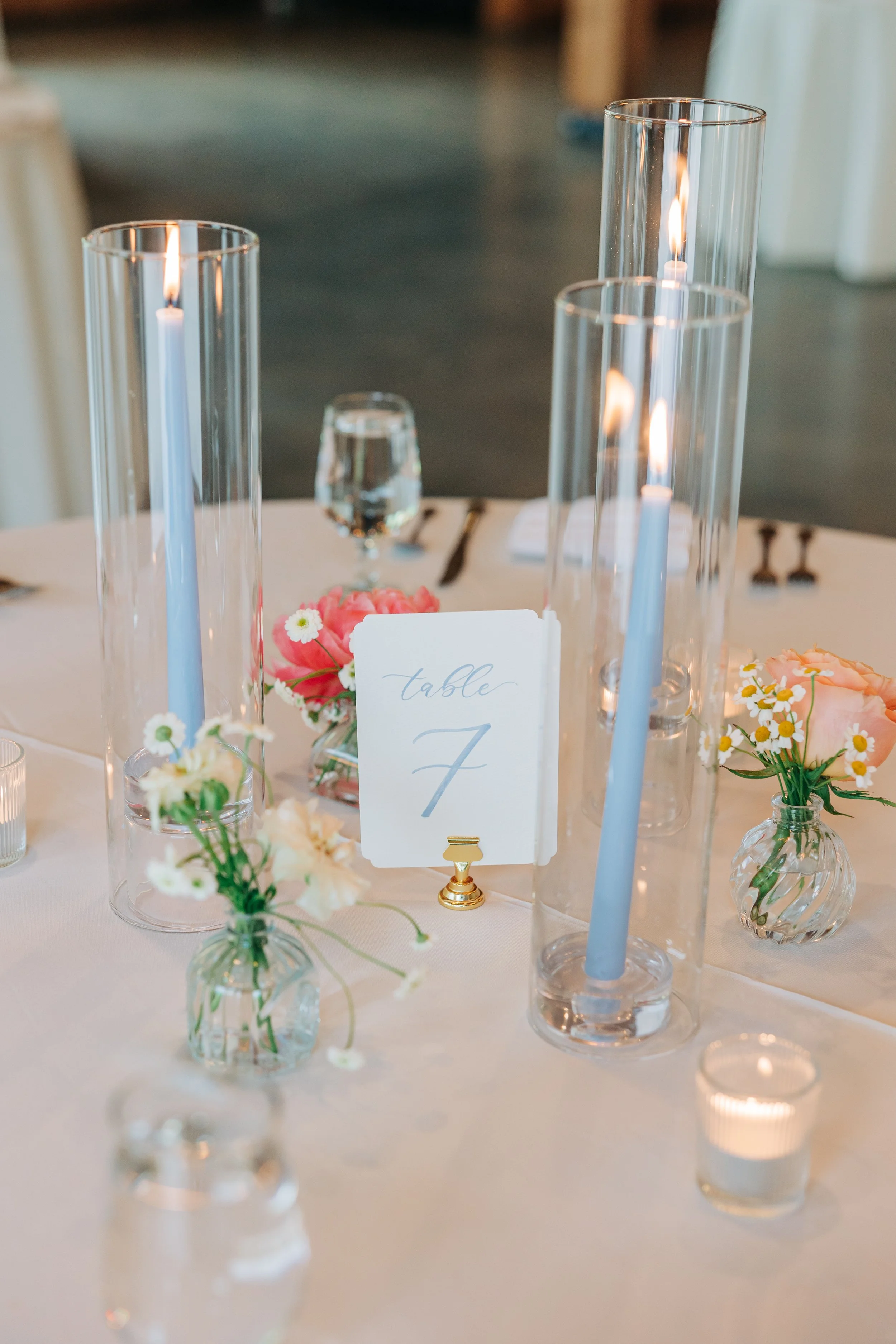 Elegant wedding reception table with glass candles, small floral arrangements, and a table number card.