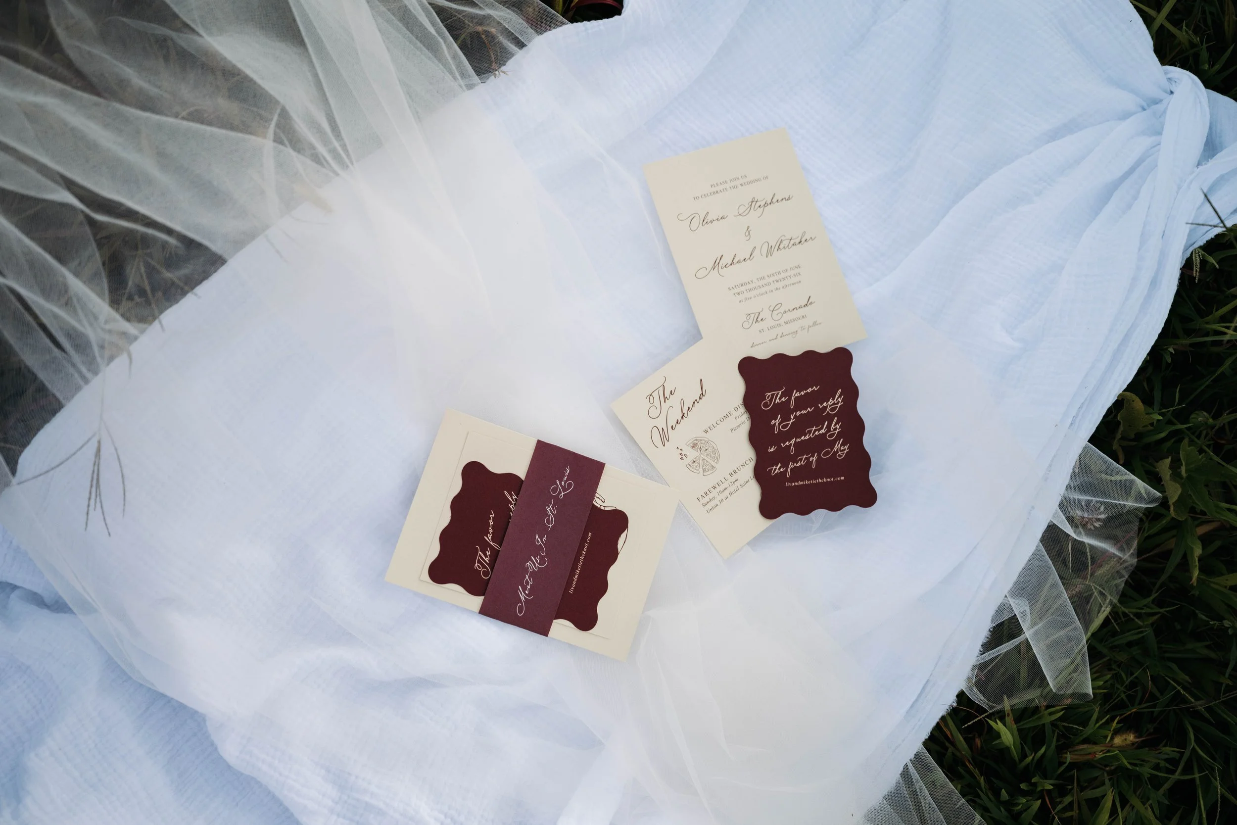 Wedding invitation and related stationery laid out on white fabric, possibly part of a wedding setup. The stationery includes a cream-colored invite with elegant script and maroon accents, along with additional cards or tags.