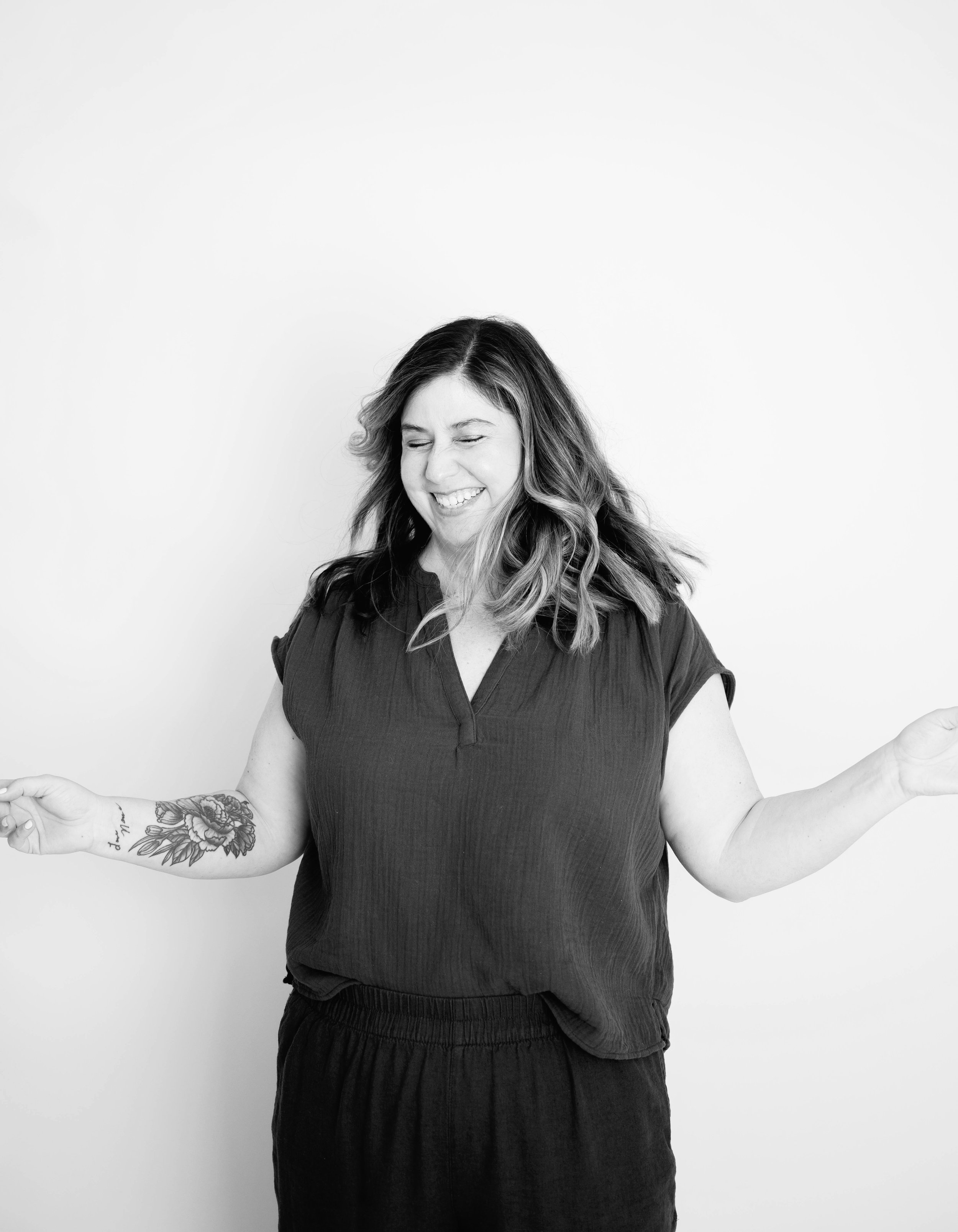 Black and white photo of a smiling woman with wavy hair, wearing a short-sleeved top, holding her arms out with palms up, standing against a plain white wall.