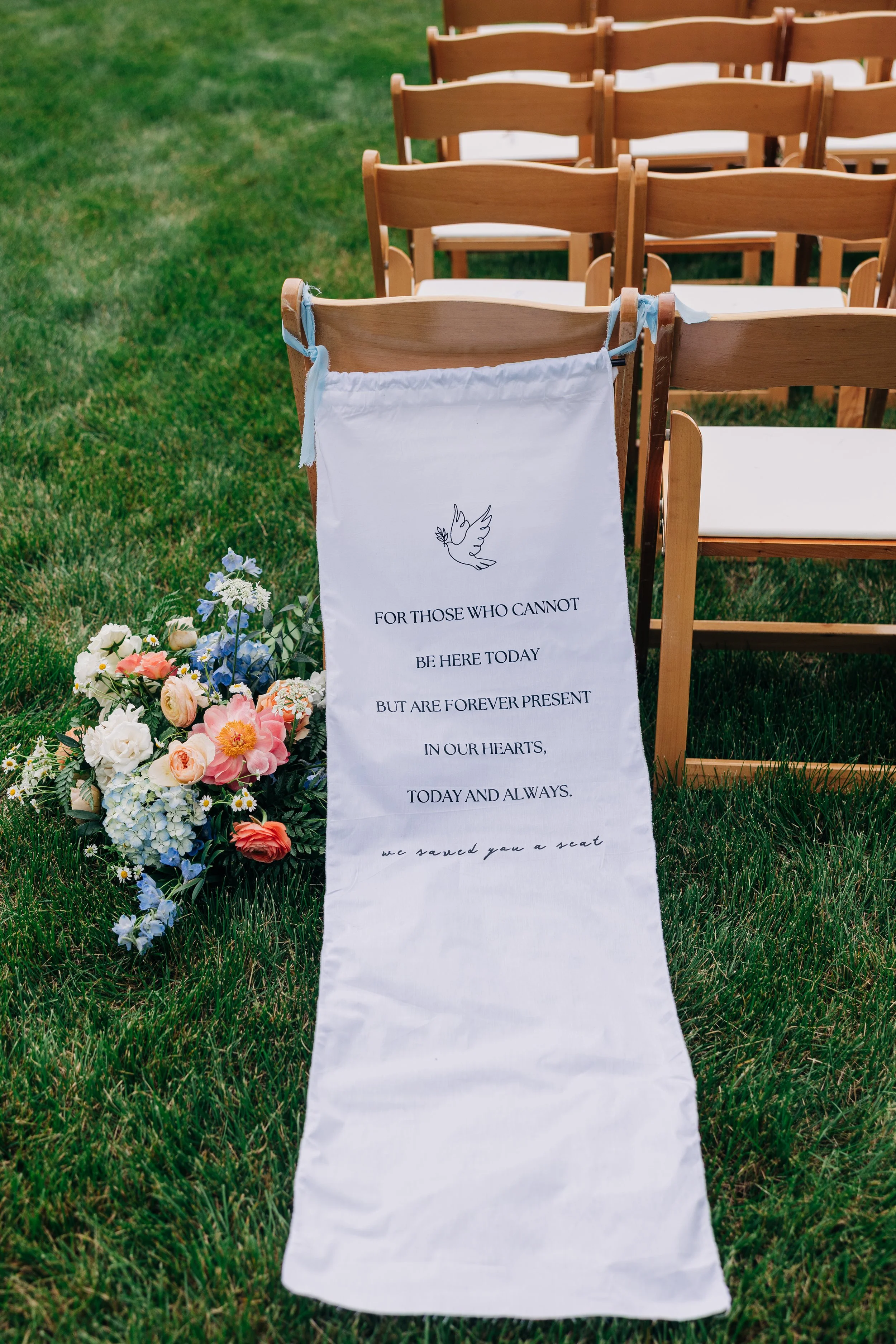 A white fabric wedding program with a dove illustration and heartfelt message, lying on a grassy area next to a bouquet of colorful flowers, with wooden chairs in the background for a wedding ceremony.