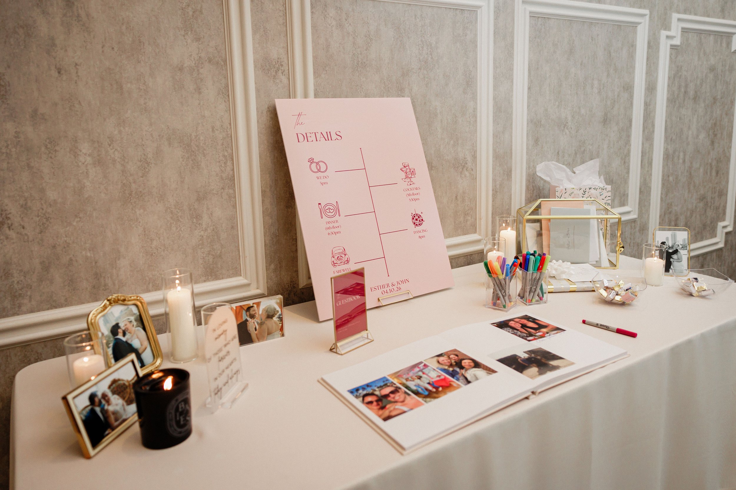 Wedding reception table decorated with framed photos, candles, a pink wedding sign, and various small items, set against a textured beige wall with decorative molding.
