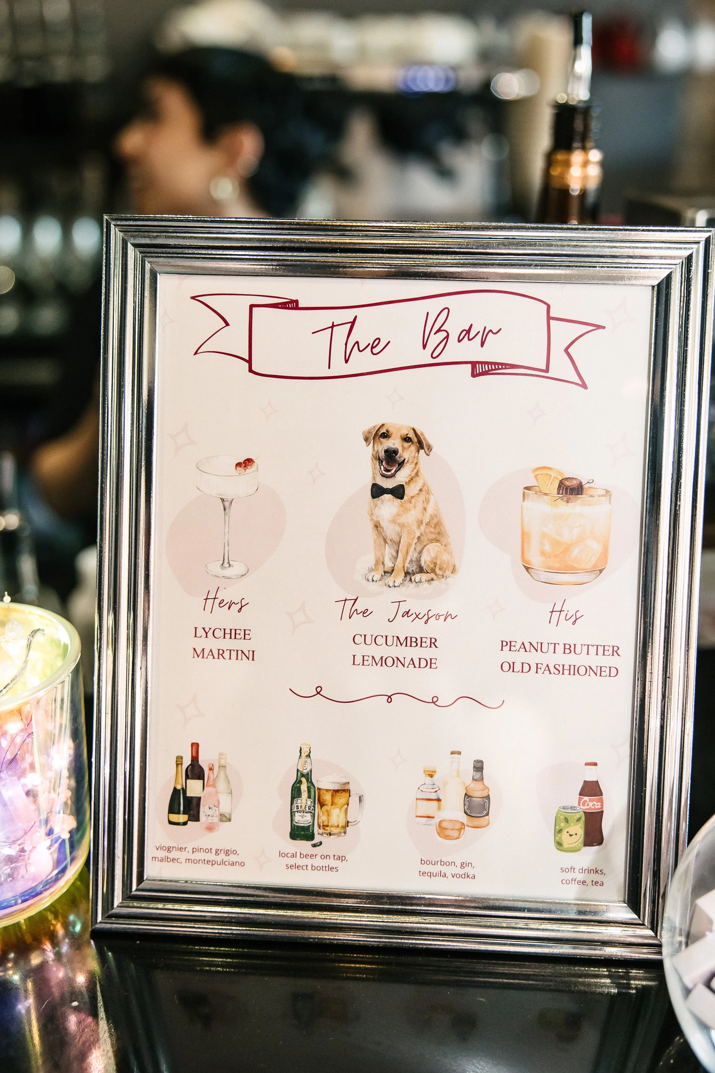 A framed menu with the title "The Bar" and three illustrations at the top: a cocktail glass labeled "Hers", a dog wearing a bowtie labeled "The Jaxson", and a glass of a cocktail labeled "His". The menu lists drink options and includes bottles of liq