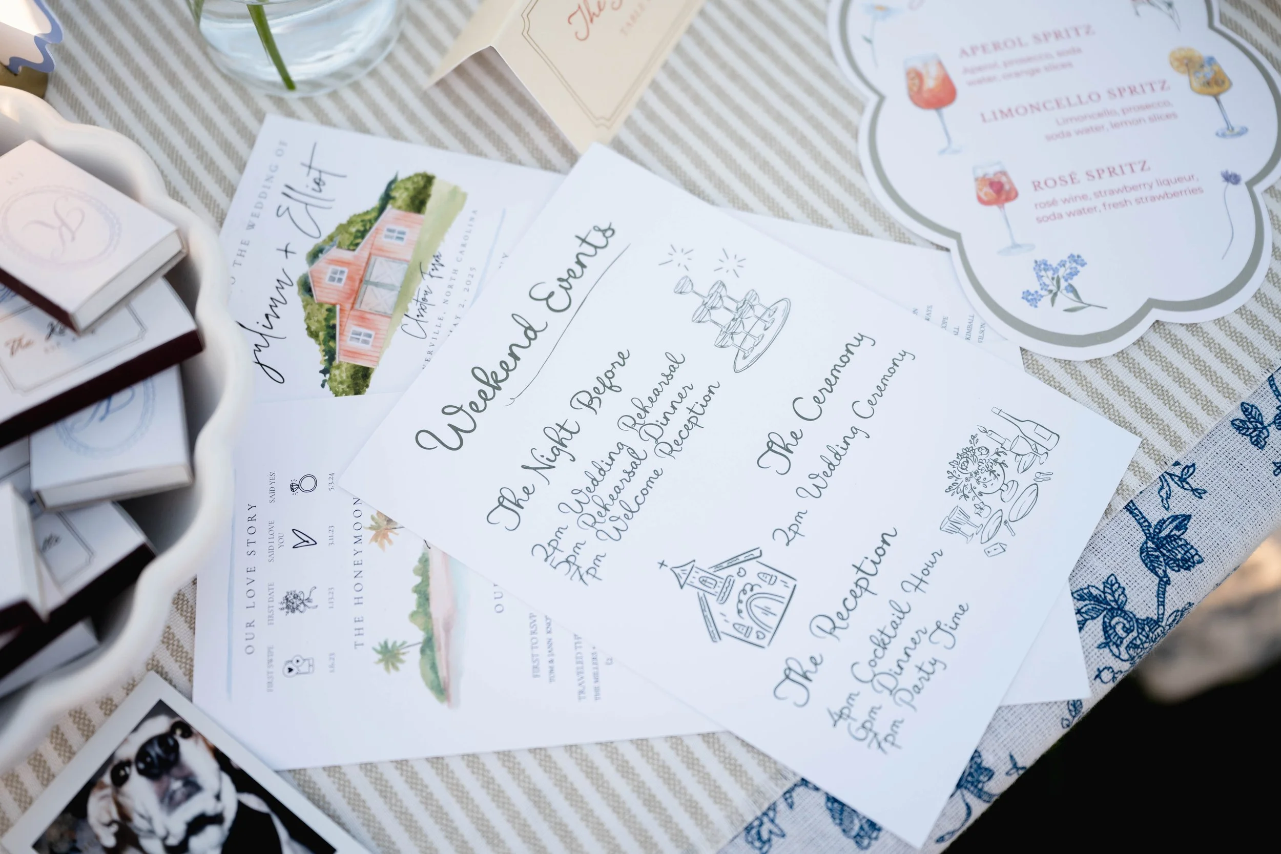 Various wedding invitations and event details printed on paper scattered on a table with a striped tablecloth, alongside a small box and a glass container with a flower.