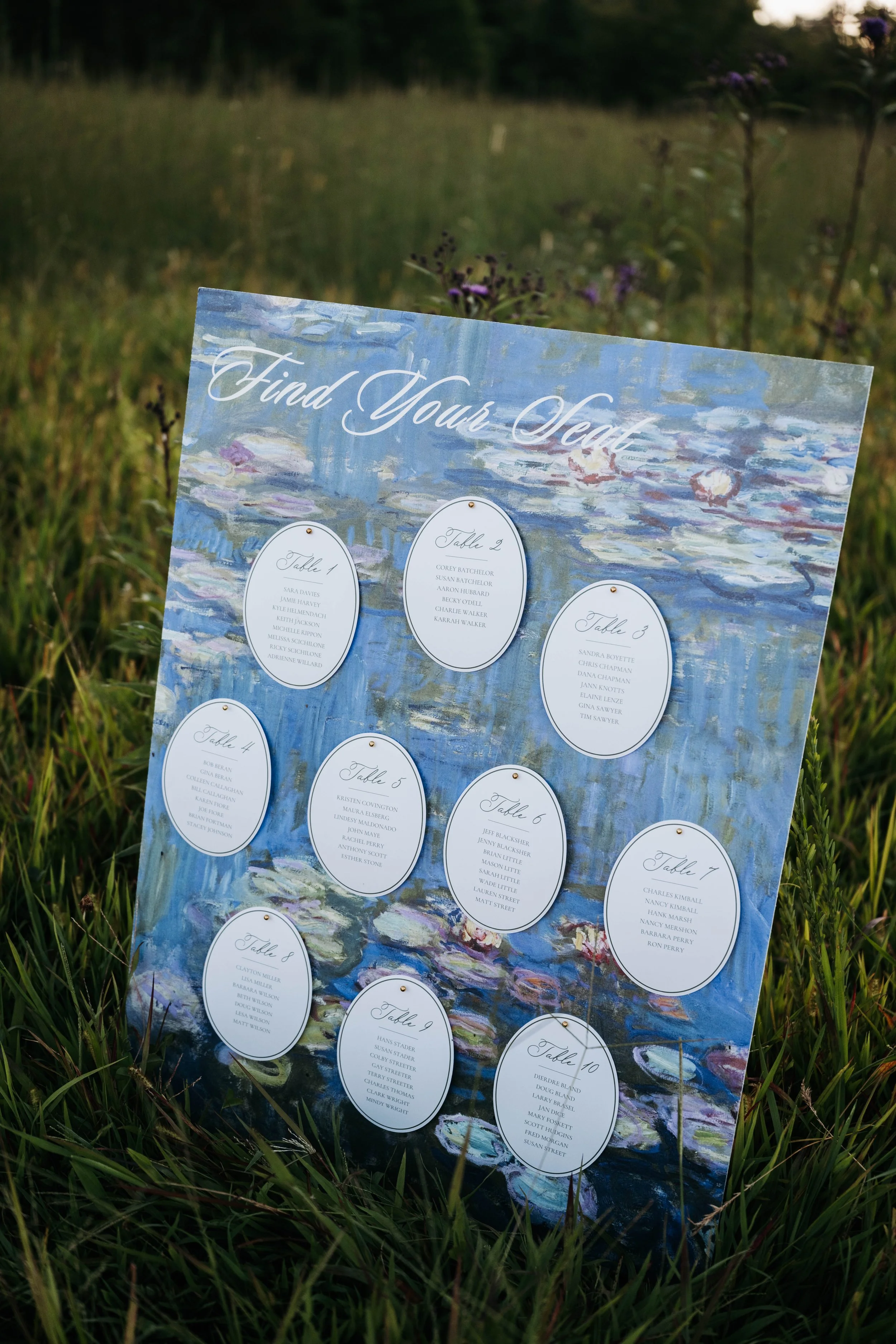 A colorful wedding seating chart displayed outdoors in a grassy area. The chart features a painting of water lilies and has tables listed with guest names underneath each table number.