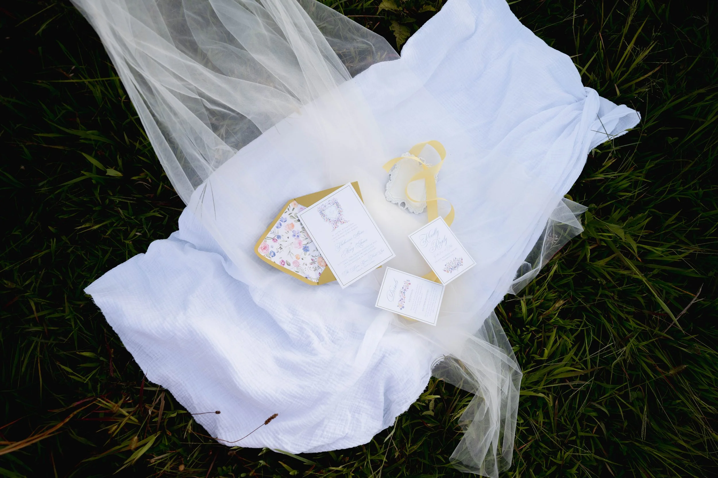Wedding invitation cards and ribbons resting on white fabric on grass with decorative veil.